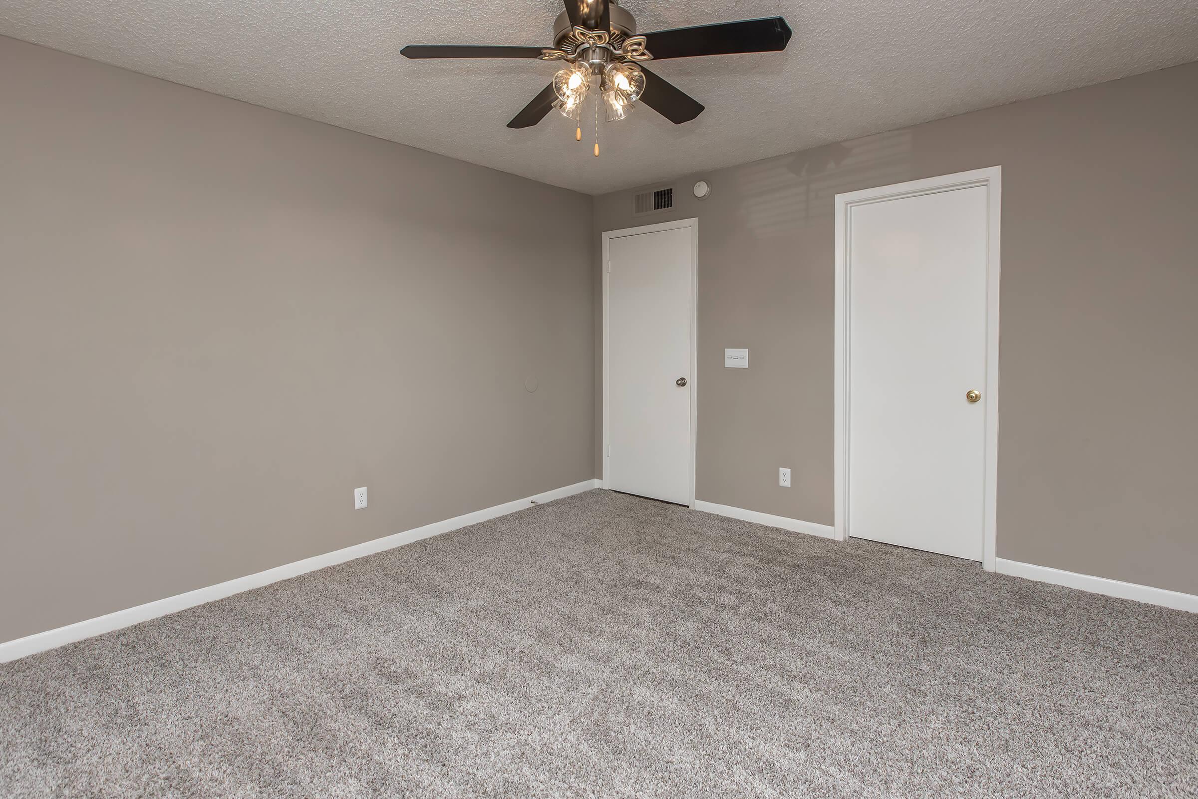A spacious, empty room featuring light gray walls and a plush carpeted floor. A ceiling fan with lights is centrally located, and there are two closed doors on one wall. The room is well-lit and has a neutral, minimalist design suitable for various purposes.