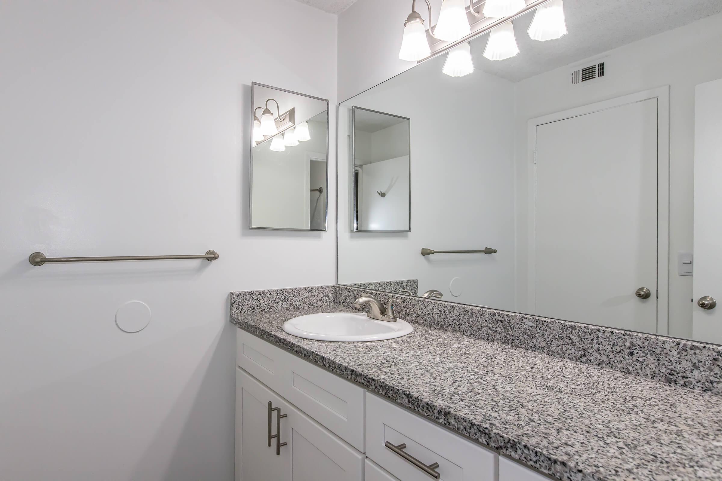 Modern bathroom featuring a white sink with a granite countertop, two mirrors above the sink, and a contemporary light fixture with multiple bulbs. The walls are painted white, and there's a towel bar installed next to the sink. A closed door is visible in the background.