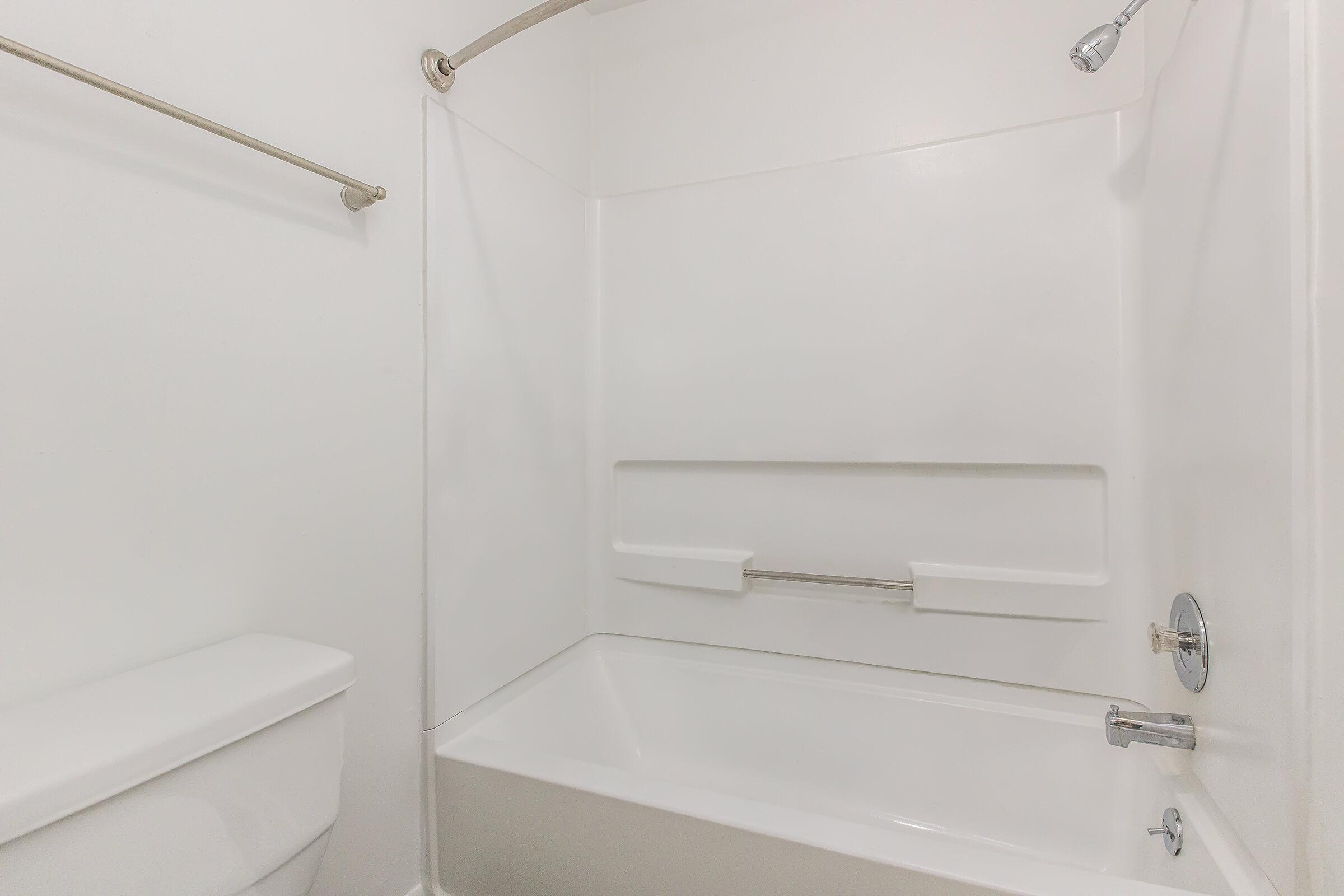A clean and minimalistic bathroom featuring a white bathtub with a chrome faucet, a showerhead above, a toilet on the left, and grab bars on the wall for accessibility. The walls are plain and white, creating a bright and spacious feel. The overall design emphasizes simplicity and functionality.