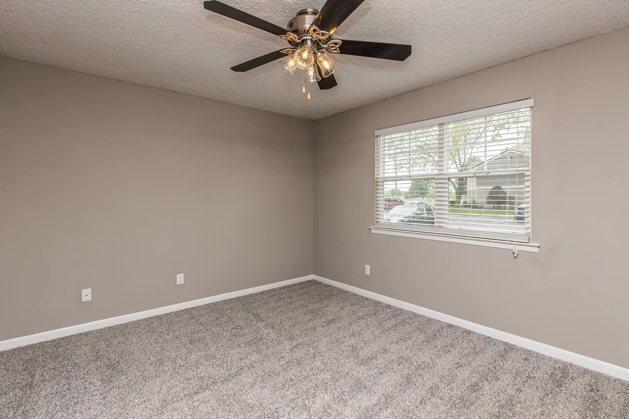 A minimalistic room featuring light gray walls and plush carpet. A ceiling fan with light fixtures is mounted on the ceiling. A window with blinds allows natural light to enter, partially revealing a view of greenery outside. The space is empty and ready for furnishing.