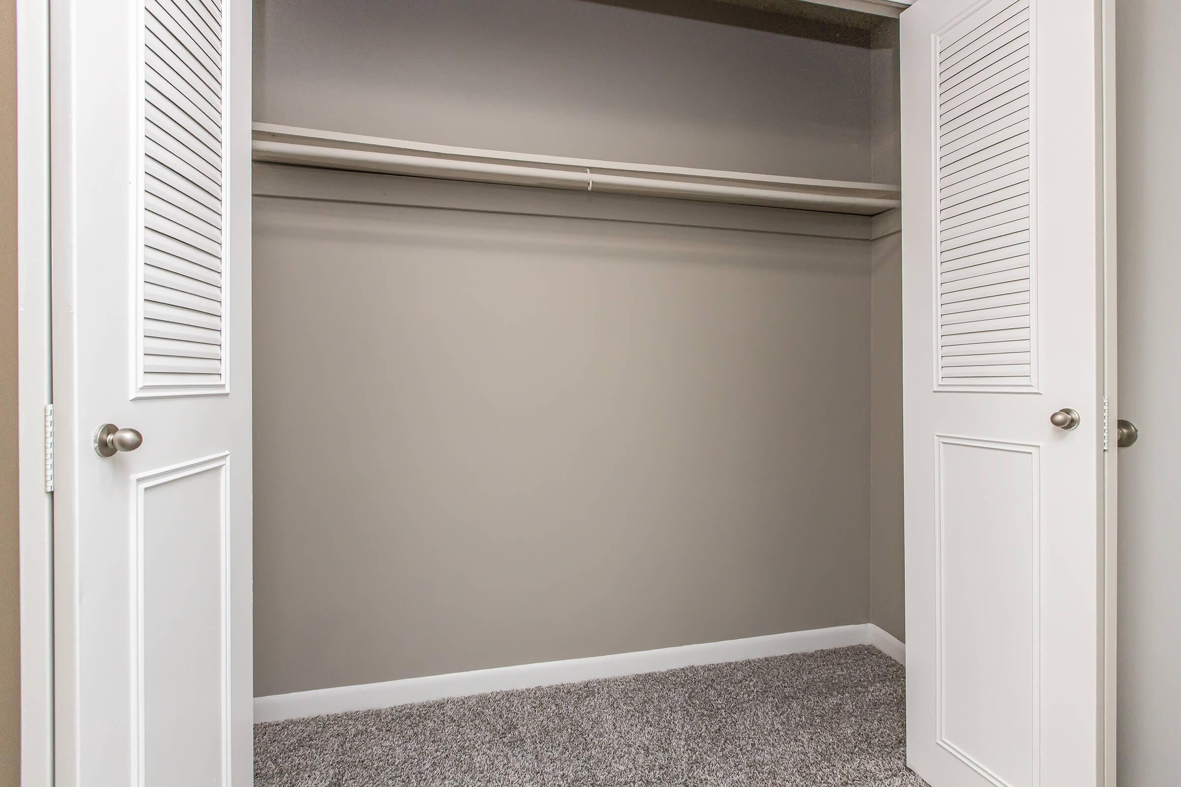 Empty closet with white bi-fold doors, featuring a single shelf and a carpeted floor. The walls are painted in a light gray color, creating a neutral and spacious appearance.