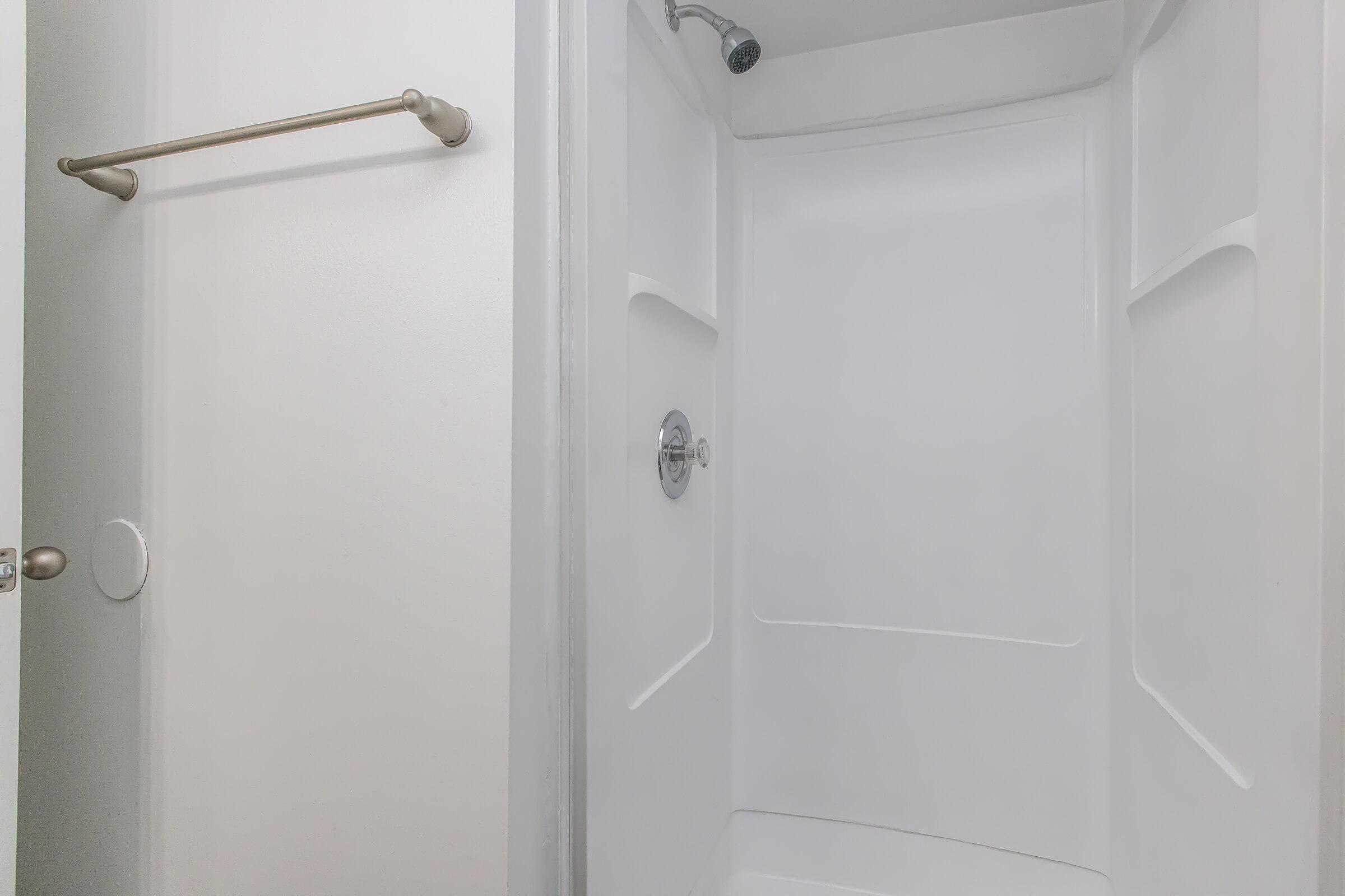 Interior view of a clean, empty shower space with a chrome showerhead and a towel bar on the wall. The walls and floor are light-colored, giving a bright and spacious appearance. The shower features a curved design with no visible fixtures or accessories inside.