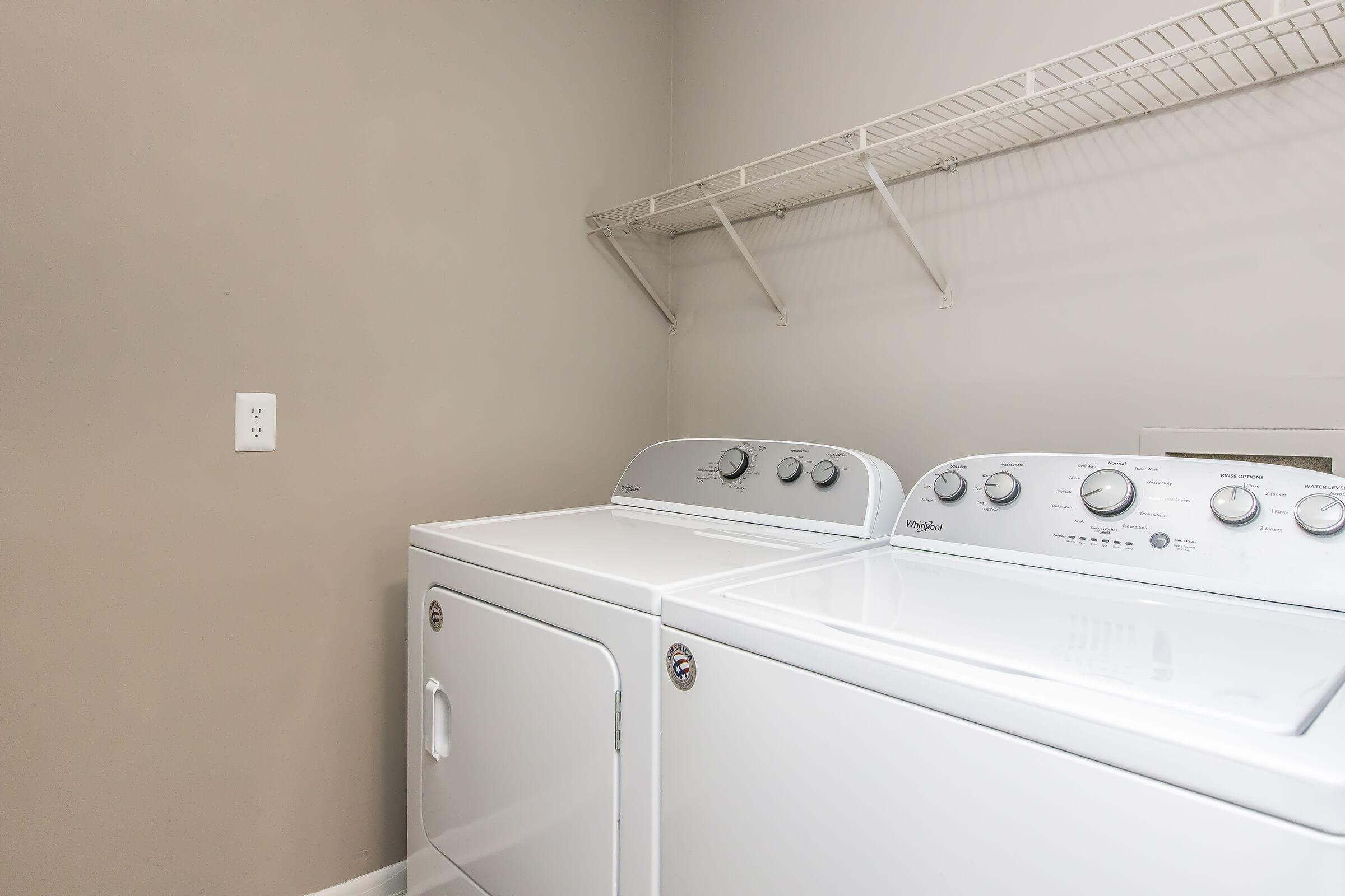 A clean laundry room featuring a white washing machine and dryer side by side. The machines have control knobs and are placed against a light gray wall. Above them, there's a wire shelf for storage. The space is well-lit and organized, emphasizing a functional area for laundry tasks.