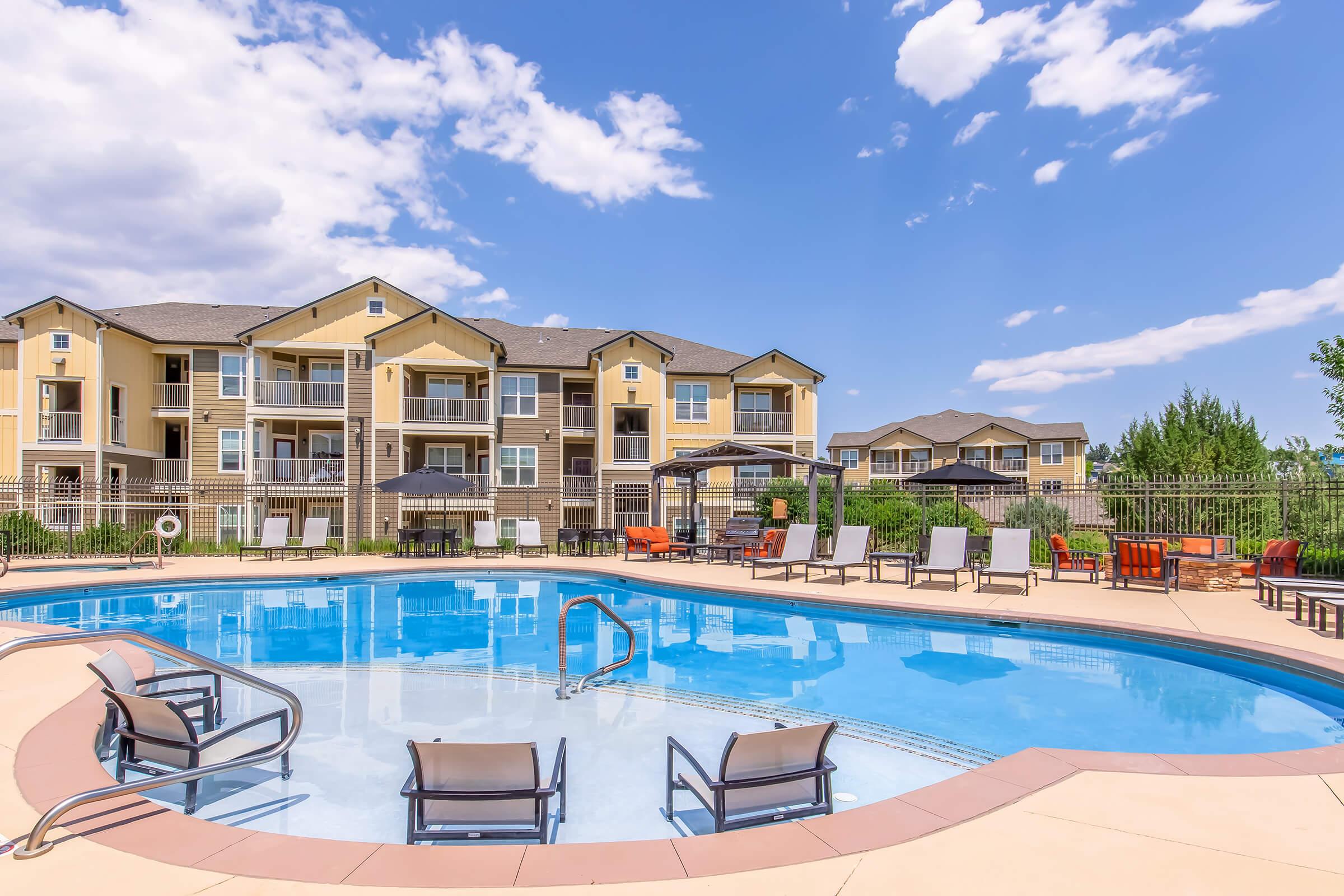 A well-maintained swimming pool surrounded by lounge chairs and patio furniture, set against a backdrop of modern apartment buildings. The scene features clear blue skies with a few clouds and greenery in the distance, evoking a relaxing outdoor atmosphere.