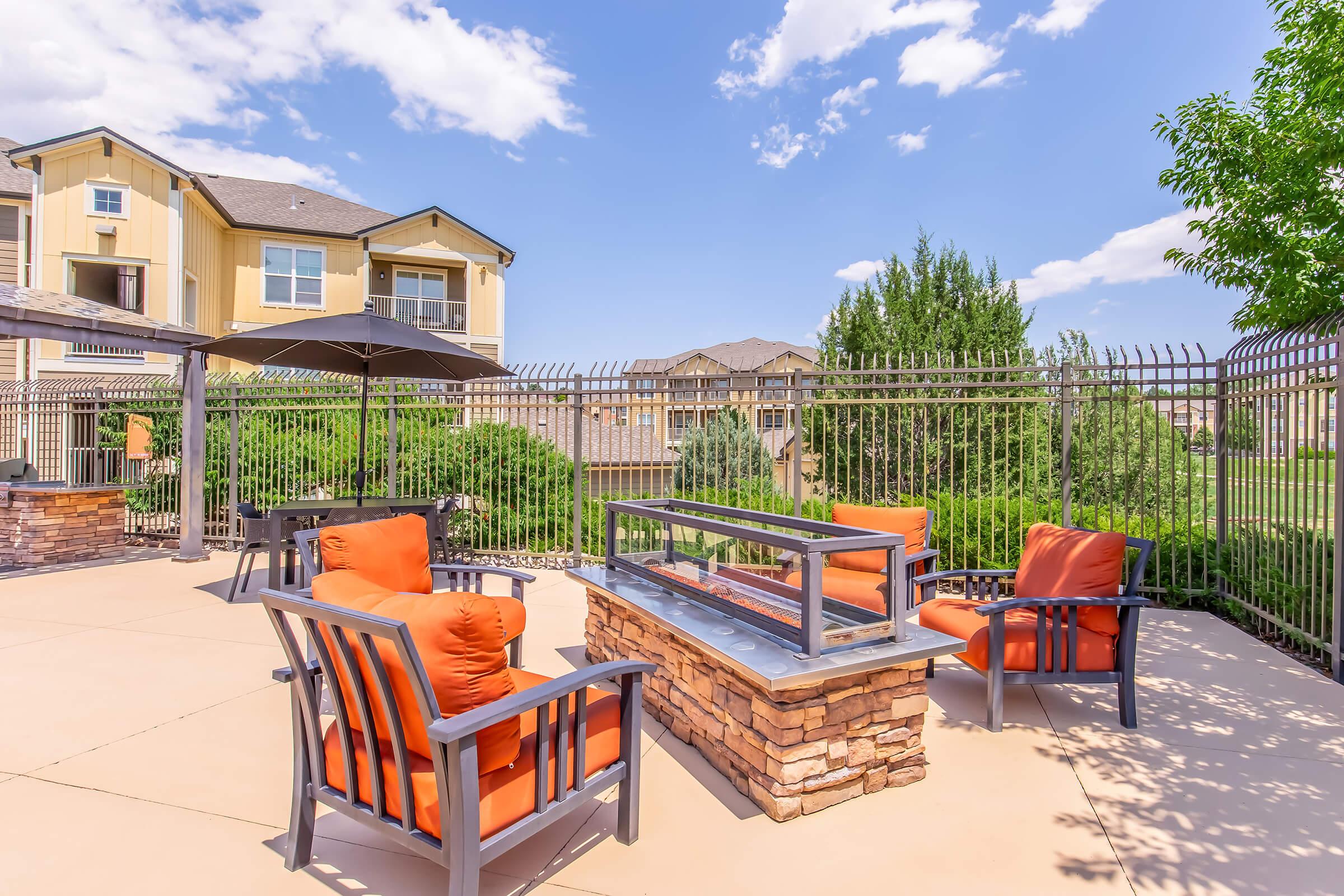 A cozy outdoor seating area featuring four orange cushioned chairs arranged around a stone fire pit, surrounded by a fenced landscape with greenery and residential buildings in the background under a bright blue sky with scattered clouds.