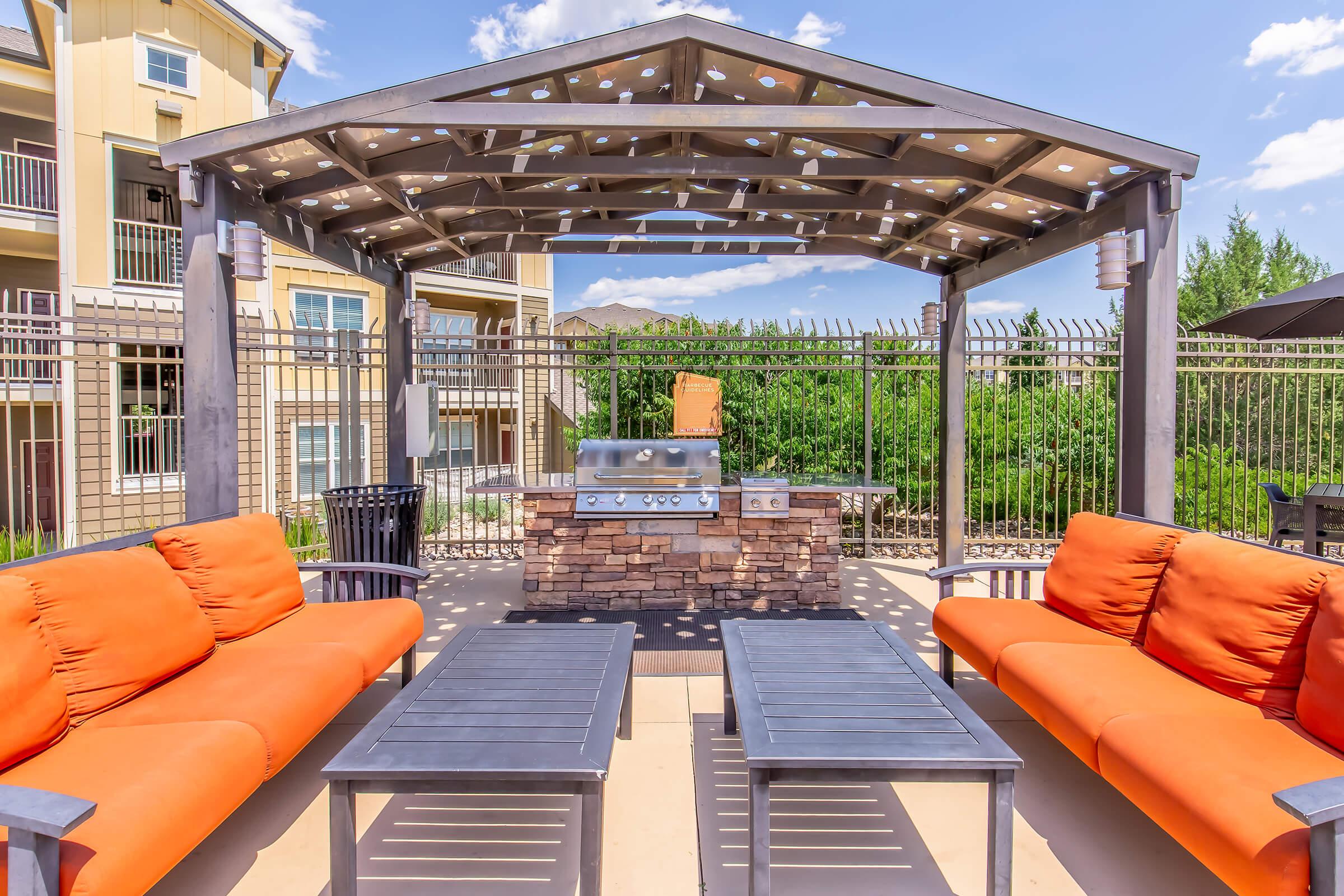A spacious outdoor patio area featuring an open gazebo with string lights, a stainless steel grill, and comfortable orange sofas with black tables. Surrounding greenery and a fenced area enhance the inviting atmosphere, ideal for gatherings and relaxation.