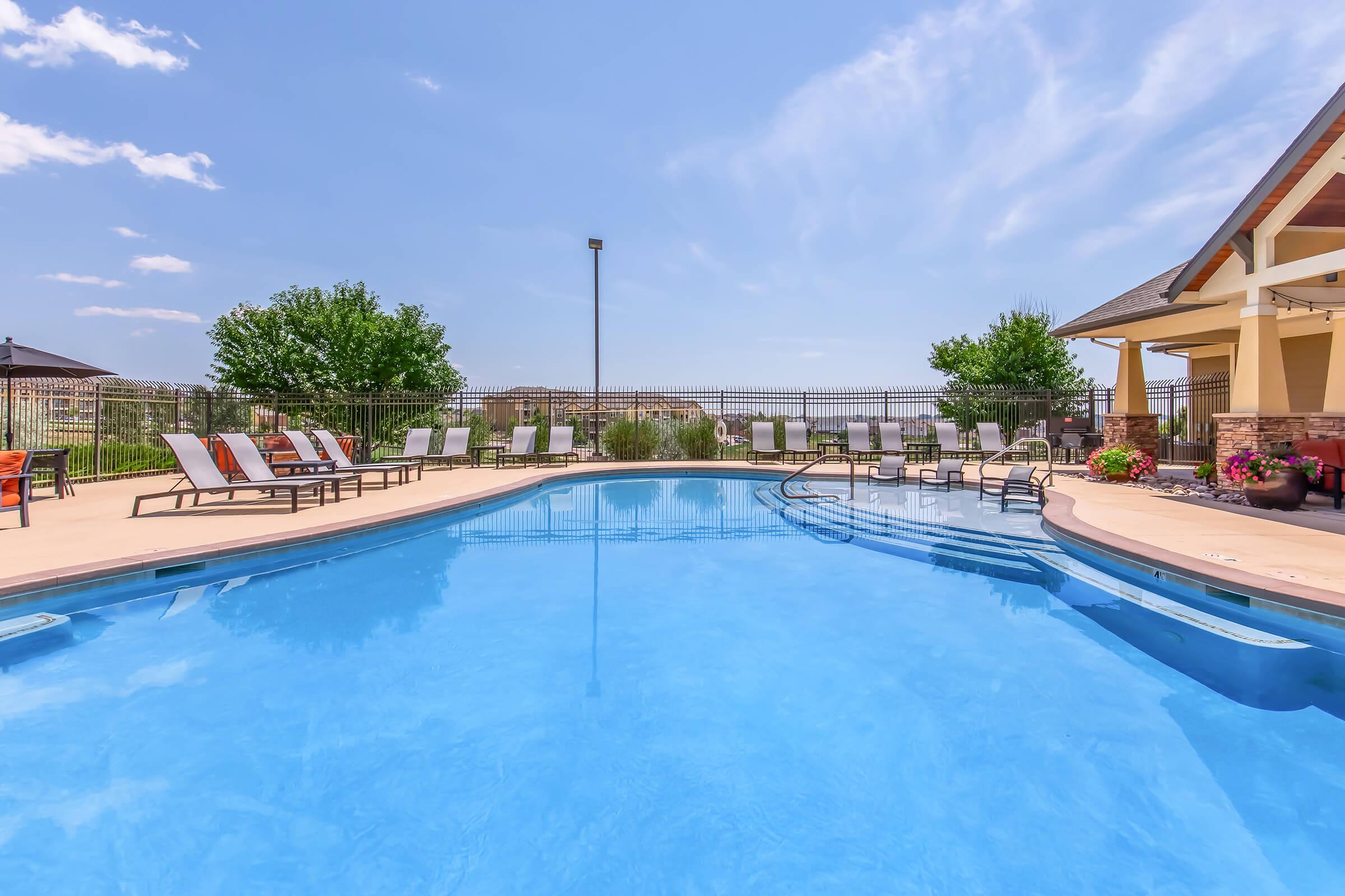 A bright blue swimming pool surrounded by lounge chairs and a well-maintained patio. Lush green trees are visible in the background, and a clear sky with fluffy clouds completes the serene outdoor setting. Ideal for relaxation and leisure.