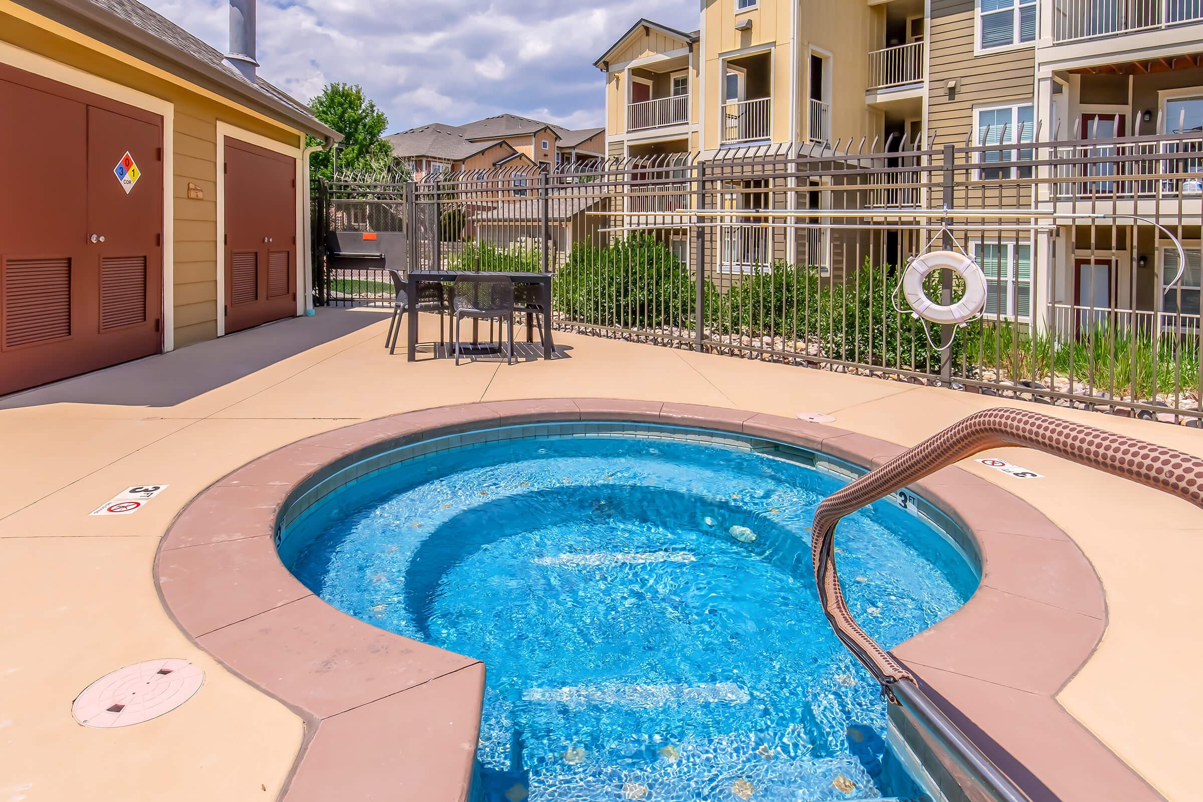 A view of a hot tub next to a swimming pool area at an apartment complex. The hot tub features steps for entry and is surrounded by a fence. In the background, there are several three-story residential buildings, grassy areas, and a cloudy sky.