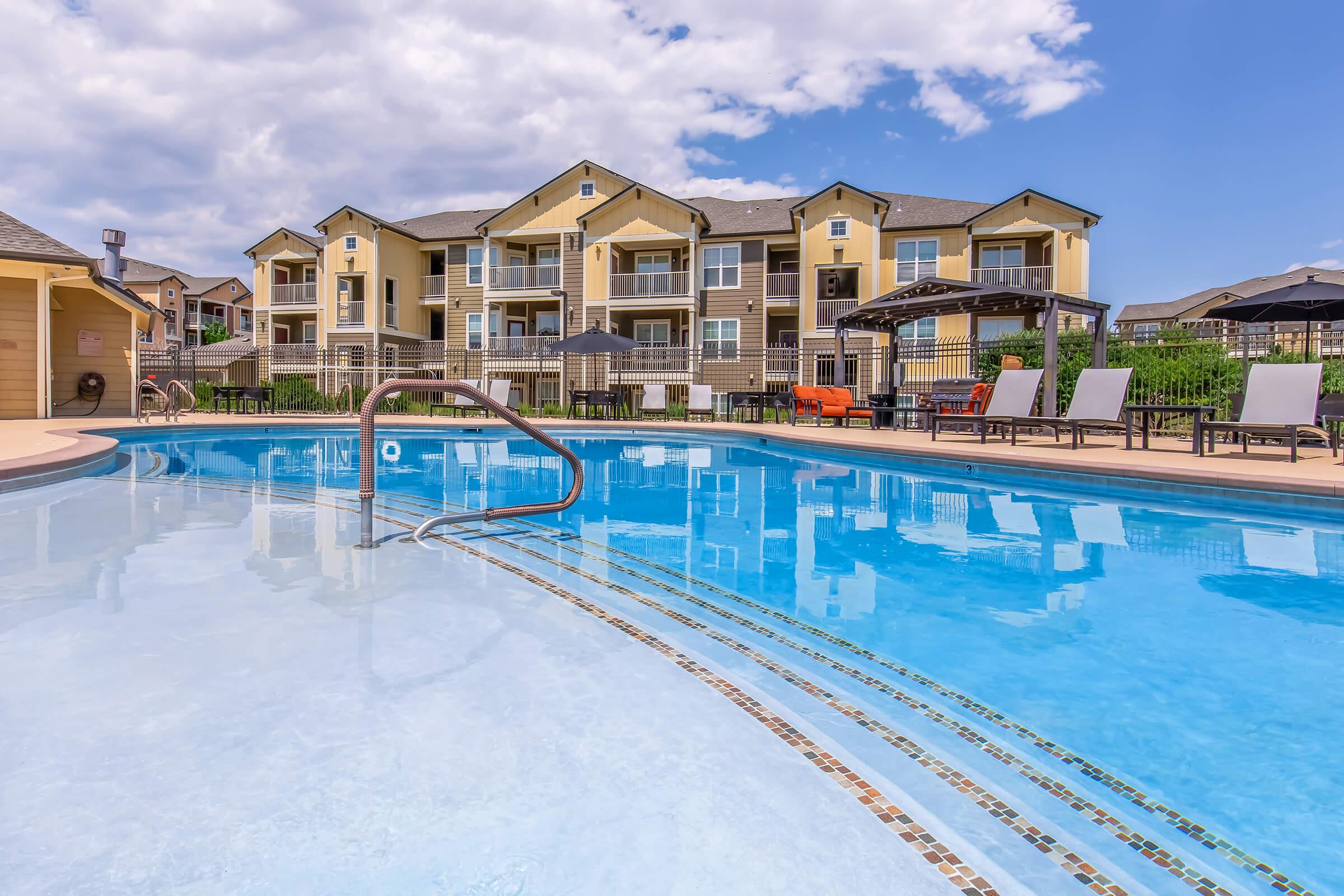 A bright outdoor pool with clear blue water, surrounded by lounge chairs and umbrellas. In the background, a multi-story apartment building with balconies overlooks the pool area under a partly cloudy sky.
