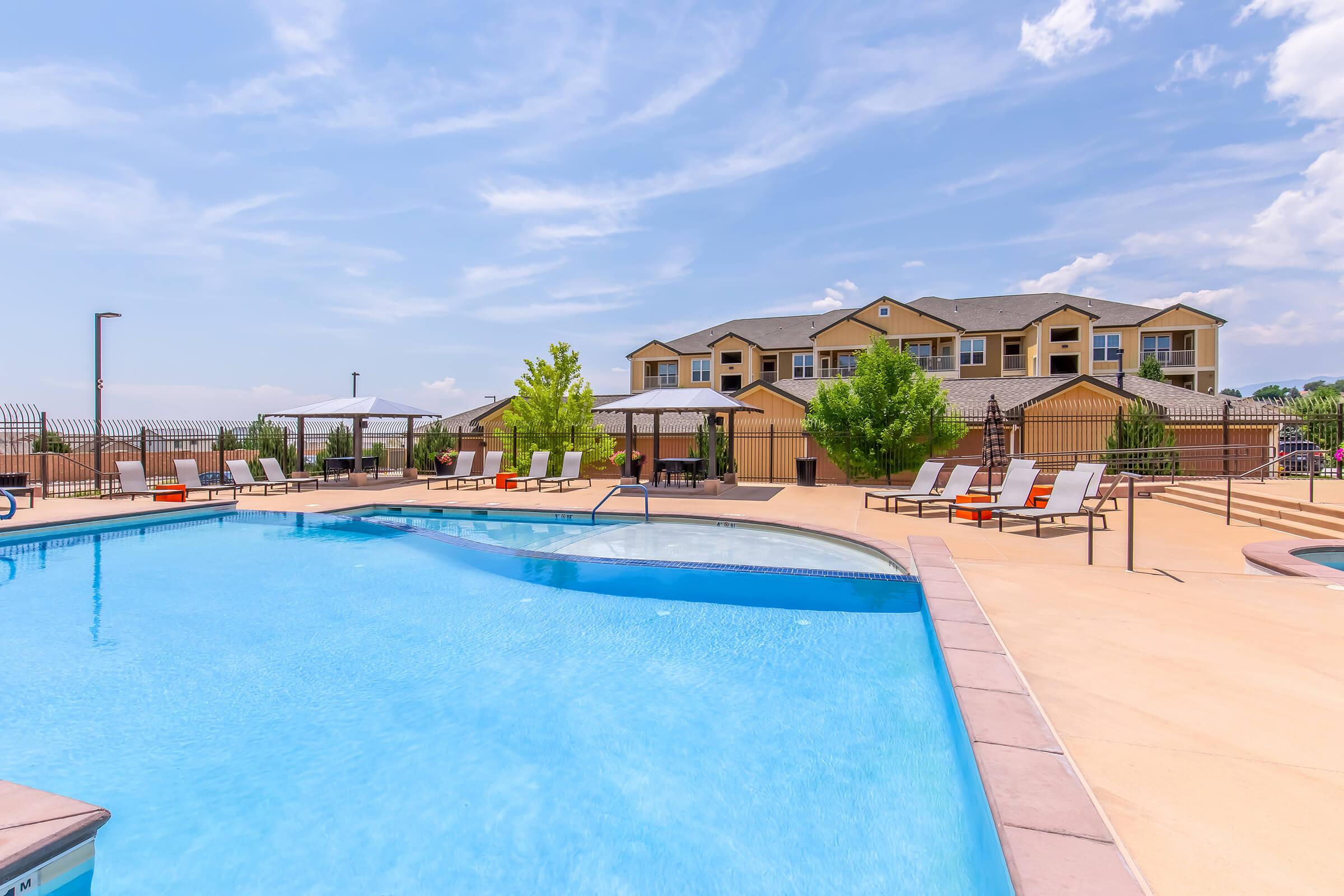 A clear blue swimming pool surrounded by lounge chairs and shaded areas, with a residential building visible in the background. The sky is bright with a few clouds, creating a relaxing outdoor atmosphere.