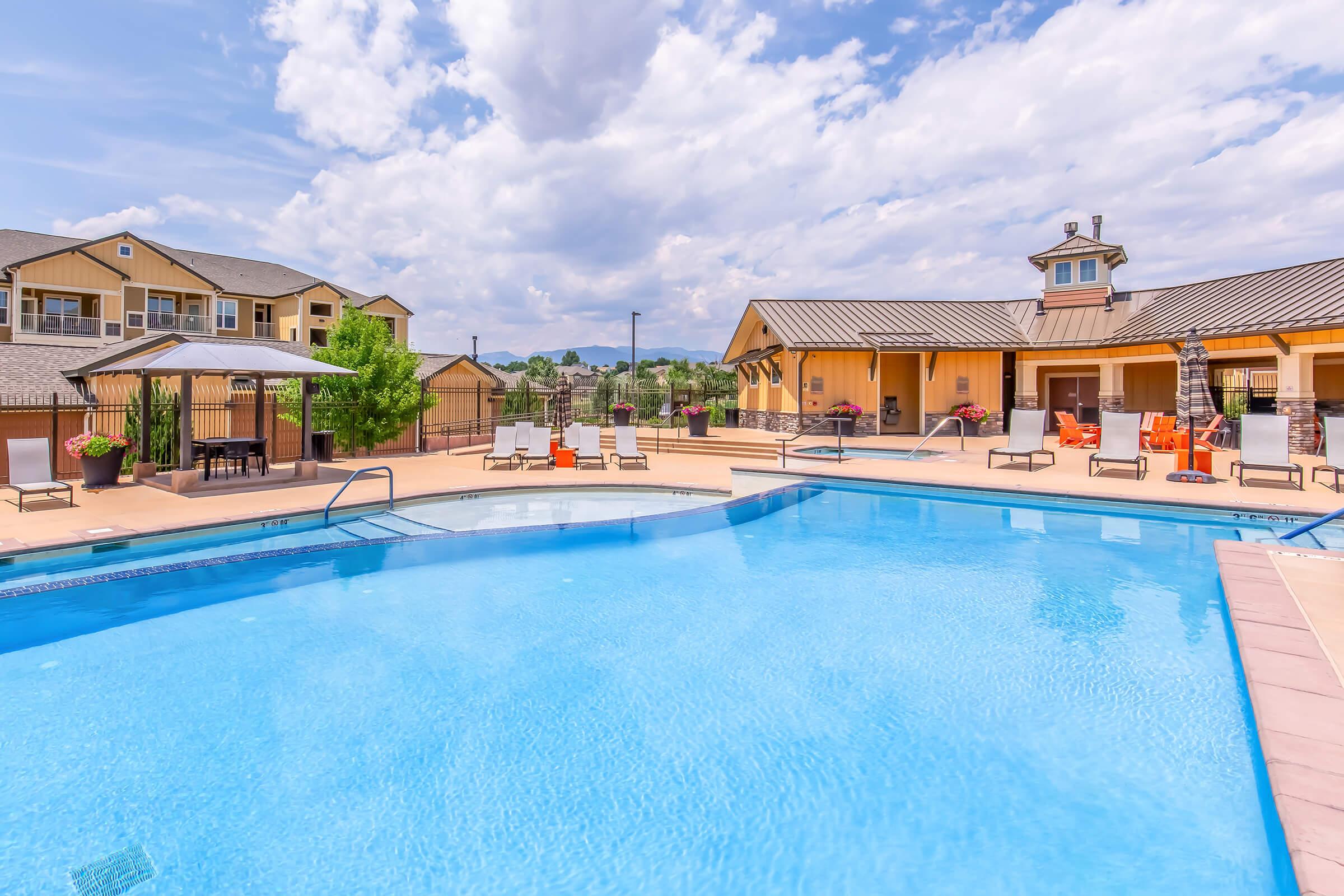 A bright and inviting swimming pool surrounded by lounge chairs and shaded areas. Nearby, a well-maintained clubhouse with a stylish architecture is visible. The sky is partly cloudy, and there are landscaped greenery and mountains in the background, creating a tranquil setting for relaxation.