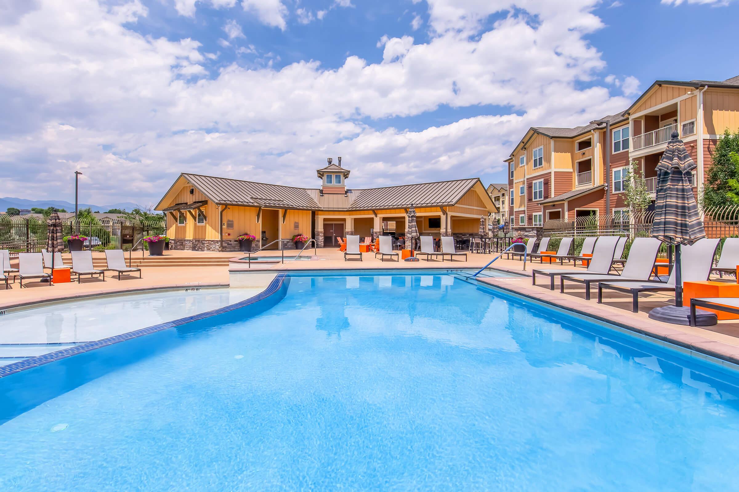 A tranquil pool area features blue water, surrounded by lounge chairs and an inviting clubhouse in the background. The scene is set against a partly cloudy sky, with well-maintained landscaping enhancing the relaxing atmosphere. Perfect for leisure and relaxation on a sunny day.