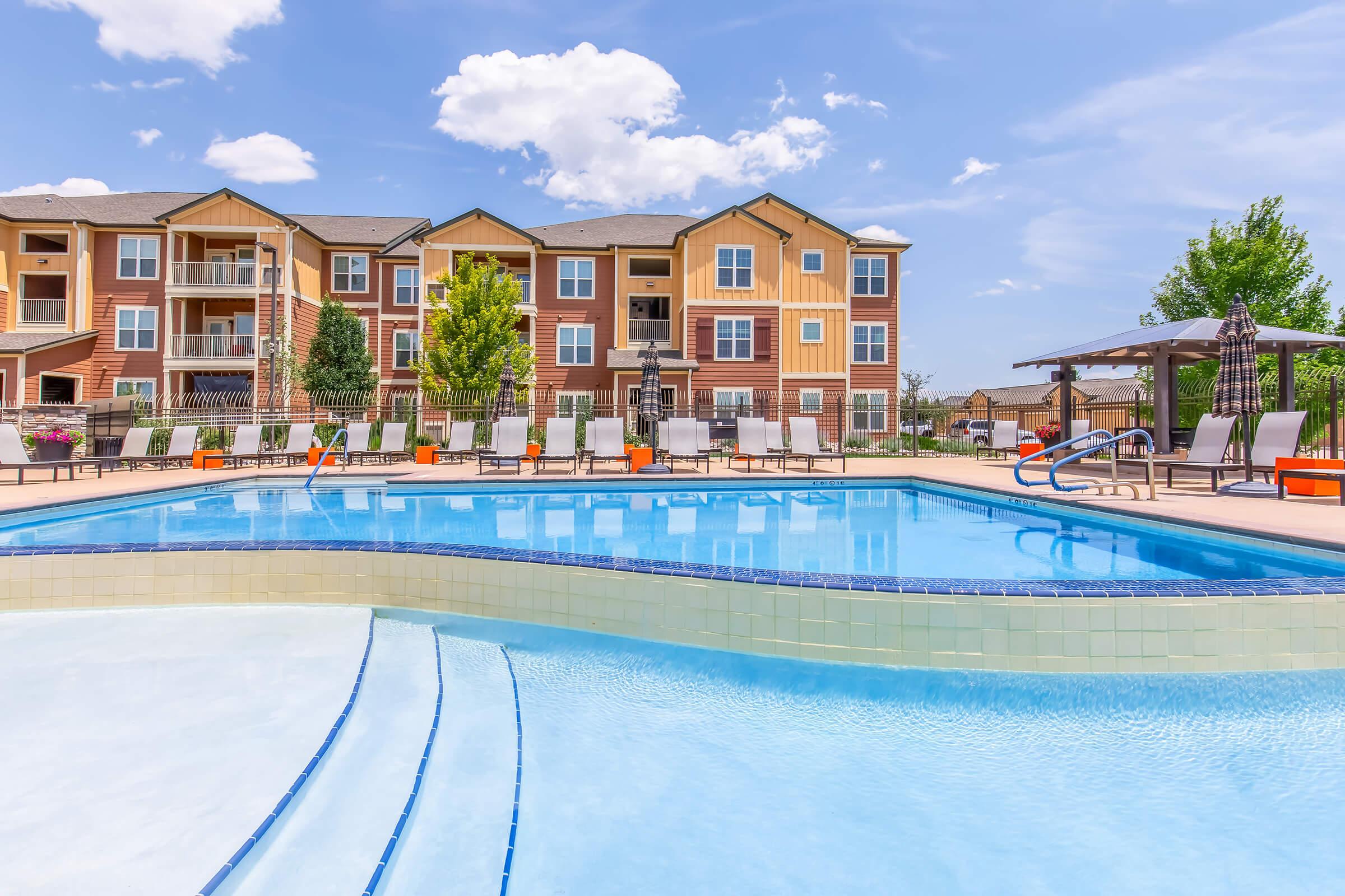 A vibrant outdoor swimming pool surrounded by lounge chairs and umbrellas, set in front of attractive apartment buildings. Lush greenery and clear blue skies enhance the serene atmosphere, making it an inviting space for relaxation and leisure.