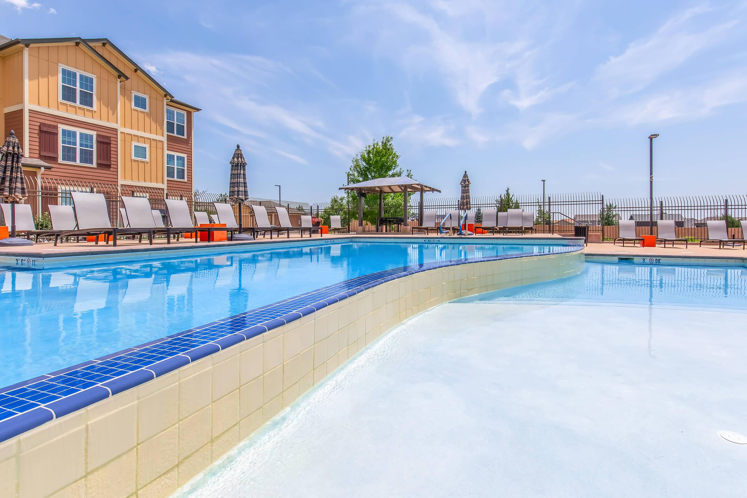 A bright outdoor swimming pool with a curved edge, surrounded by lounge chairs and umbrellas. In the background, there are several buildings and greenery under a clear blue sky. The pool area reflects a sunny and inviting atmosphere, perfect for relaxation.