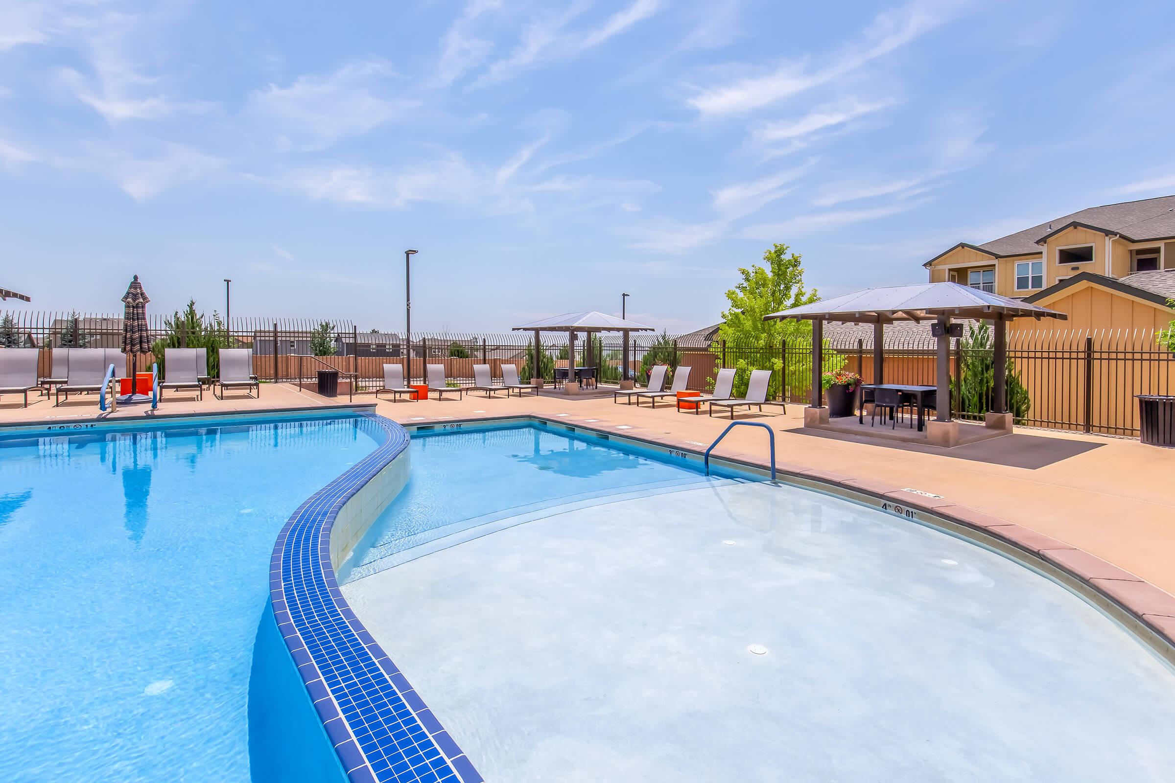 A clear blue swimming pool with a curved design, surrounded by lounge chairs and umbrellas. Nearby, there are shaded seating areas and a fence in the background. The scene is bright and sunny, with a few residential buildings visible on the right.