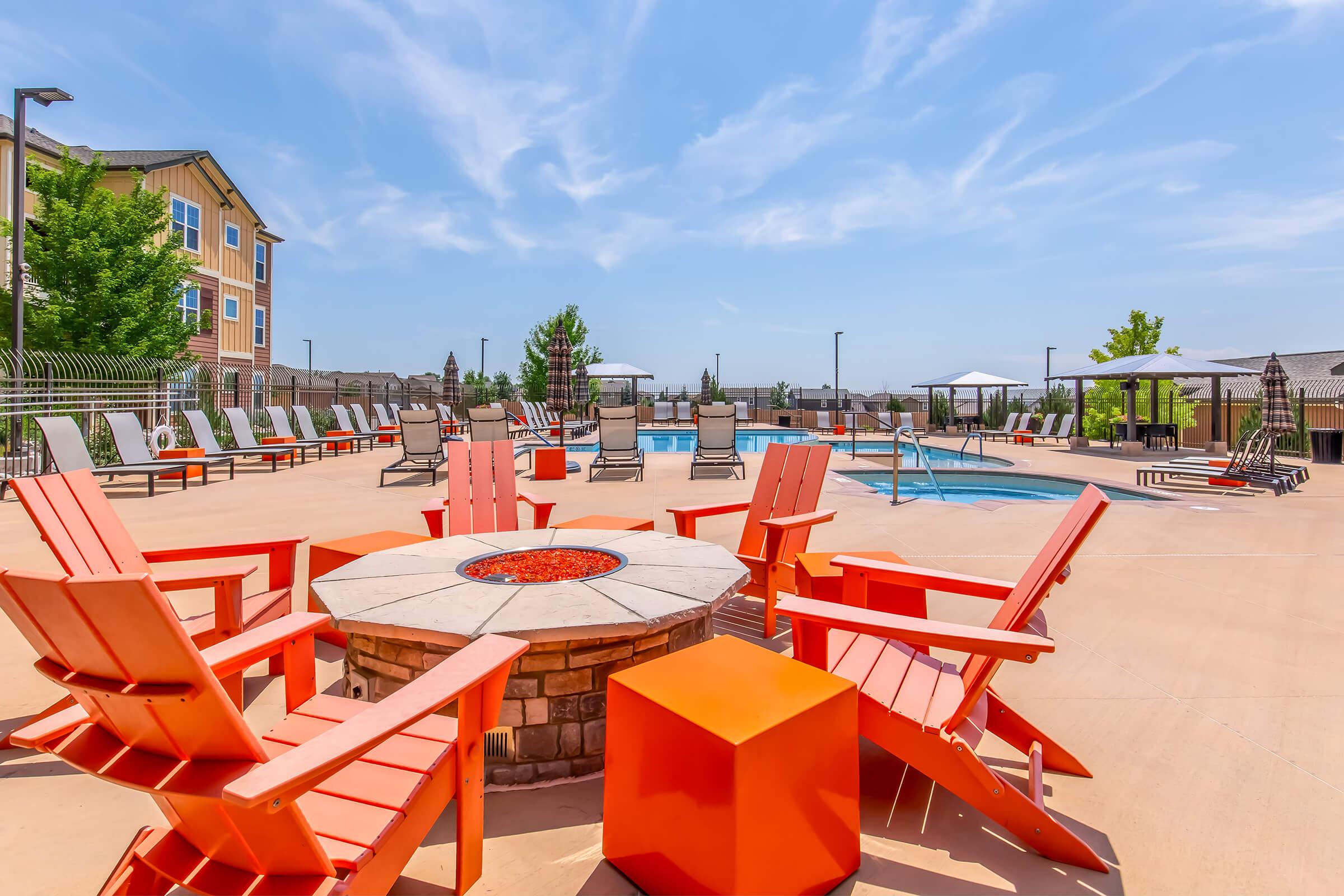 Outdoor pool area with bright orange Adirondack chairs arranged around a fire pit. Surrounding the pool are lounge chairs and shade umbrellas. Lush greenery and modern apartment buildings in the background under a blue sky with wispy clouds. Perfect for relaxation and social gatherings.
