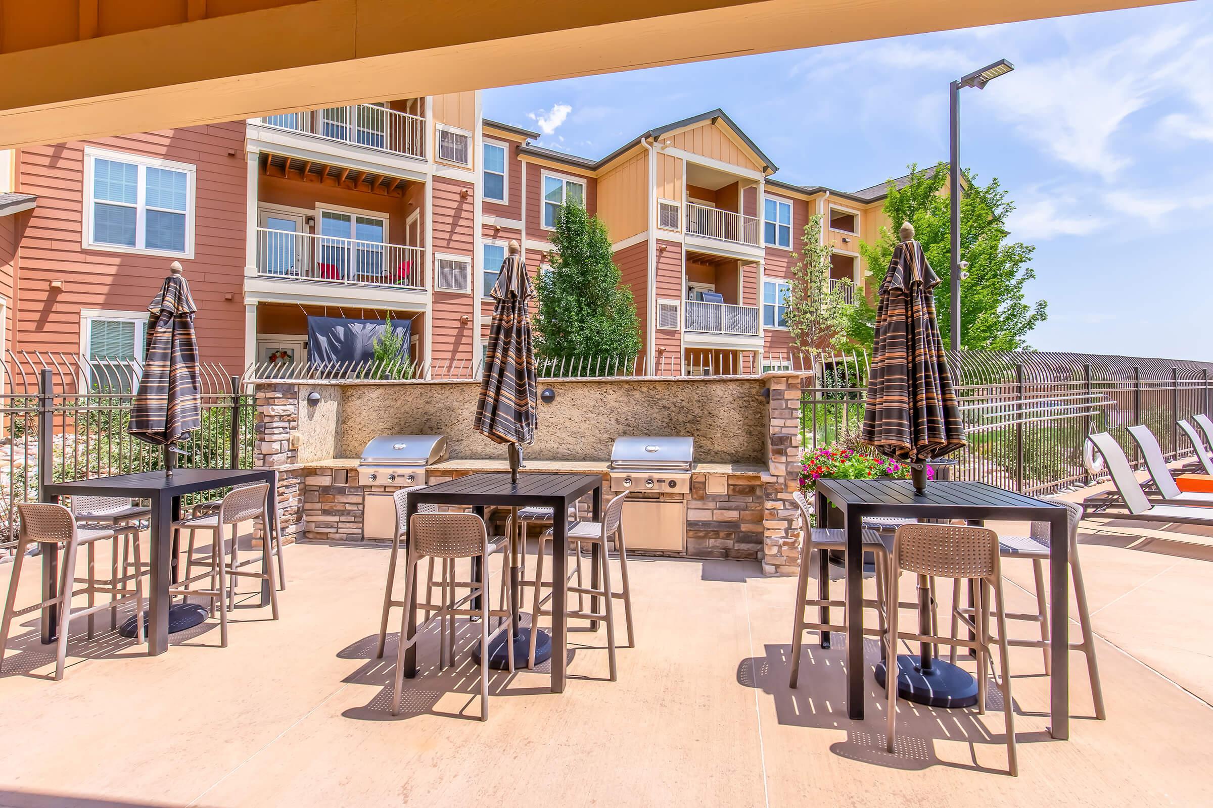 Outdoor grilling area featuring charcoal grills, tables, and chairs, with patio umbrellas. In the background, there are apartment buildings with balconies and greenery. The scene is bright and welcoming, perfect for outdoor dining and gatherings.