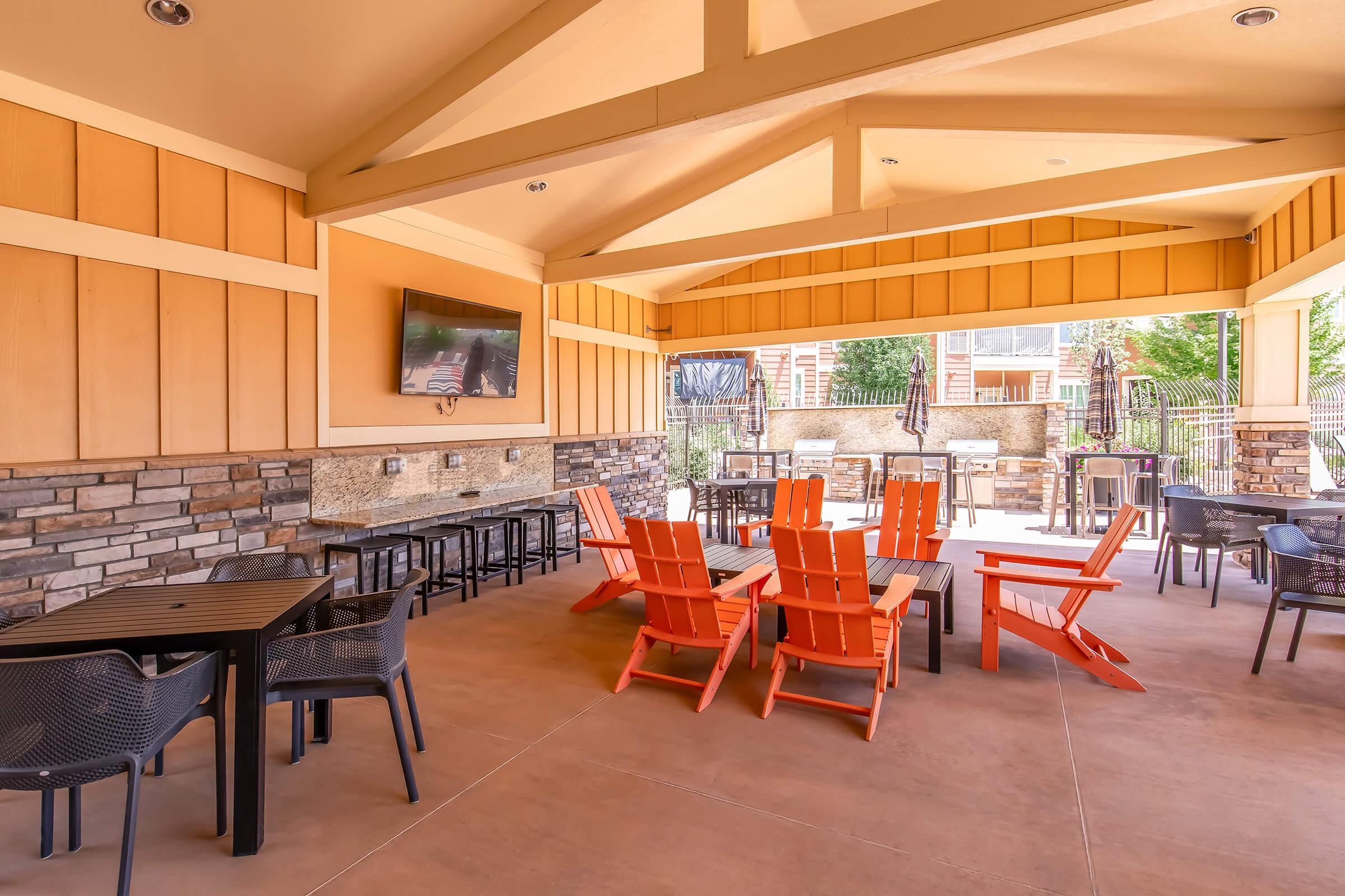 Outdoor communal space featuring a covered patio with orange adirondack chairs, black tables, and a stone accent wall. A large television is mounted on the wall, and the area is surrounded by greenery, creating a welcoming atmosphere for relaxation and socializing.