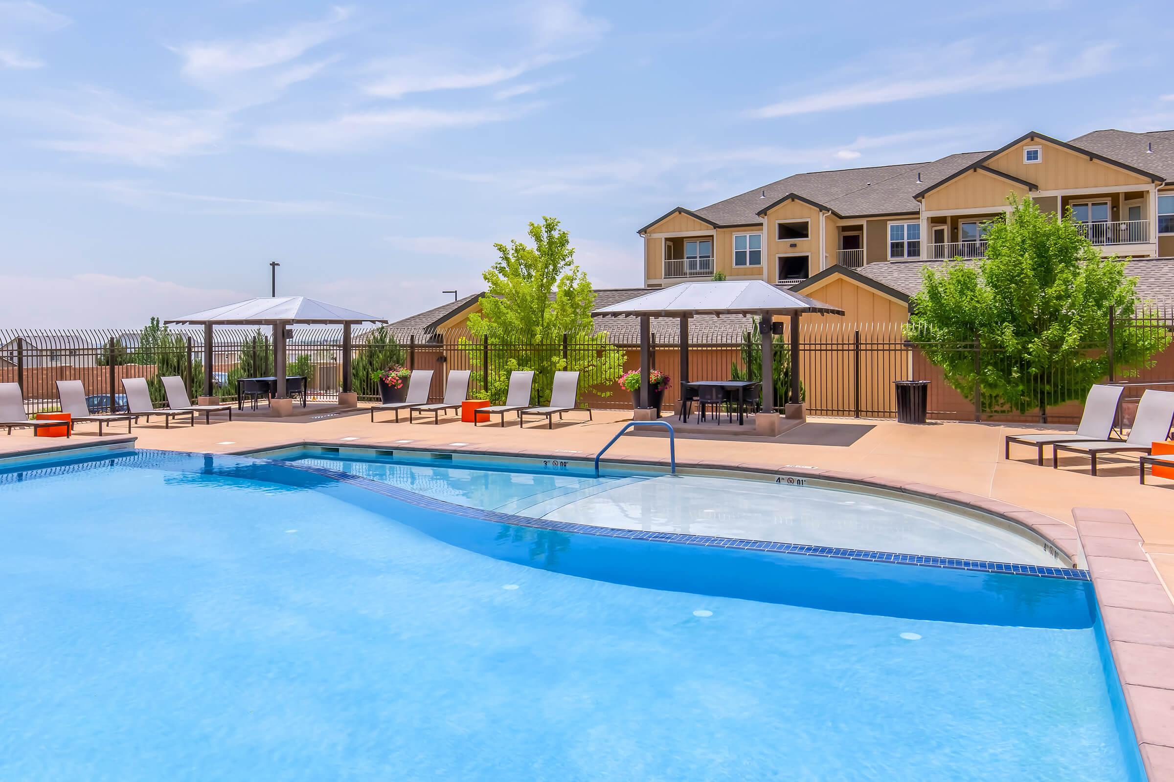 A bright outdoor swimming pool with blue water, surrounded by lounge chairs and shaded cabanas. Lush green trees and a secure fence border the area, while an apartment building is visible in the background under a clear blue sky. The scene conveys a relaxed and inviting atmosphere.