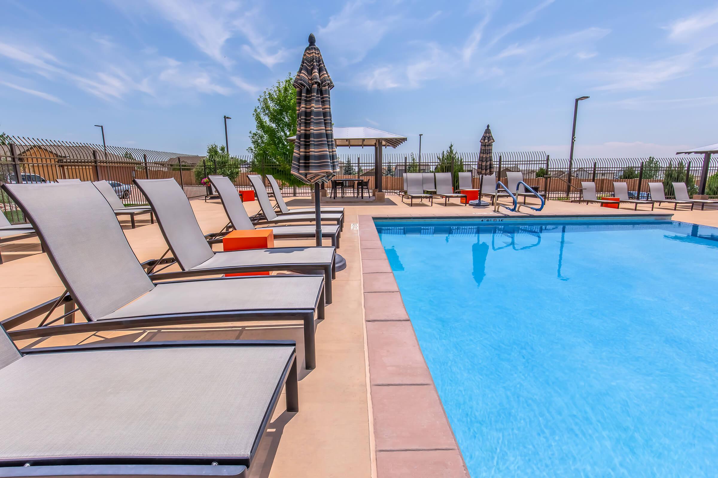 A bright and inviting outdoor swimming pool area featuring several lounge chairs lined up around the pool. An umbrella is placed near the pool, and a shaded seating area is visible in the background. The surroundings are well-maintained with greenery and a clear blue sky above.