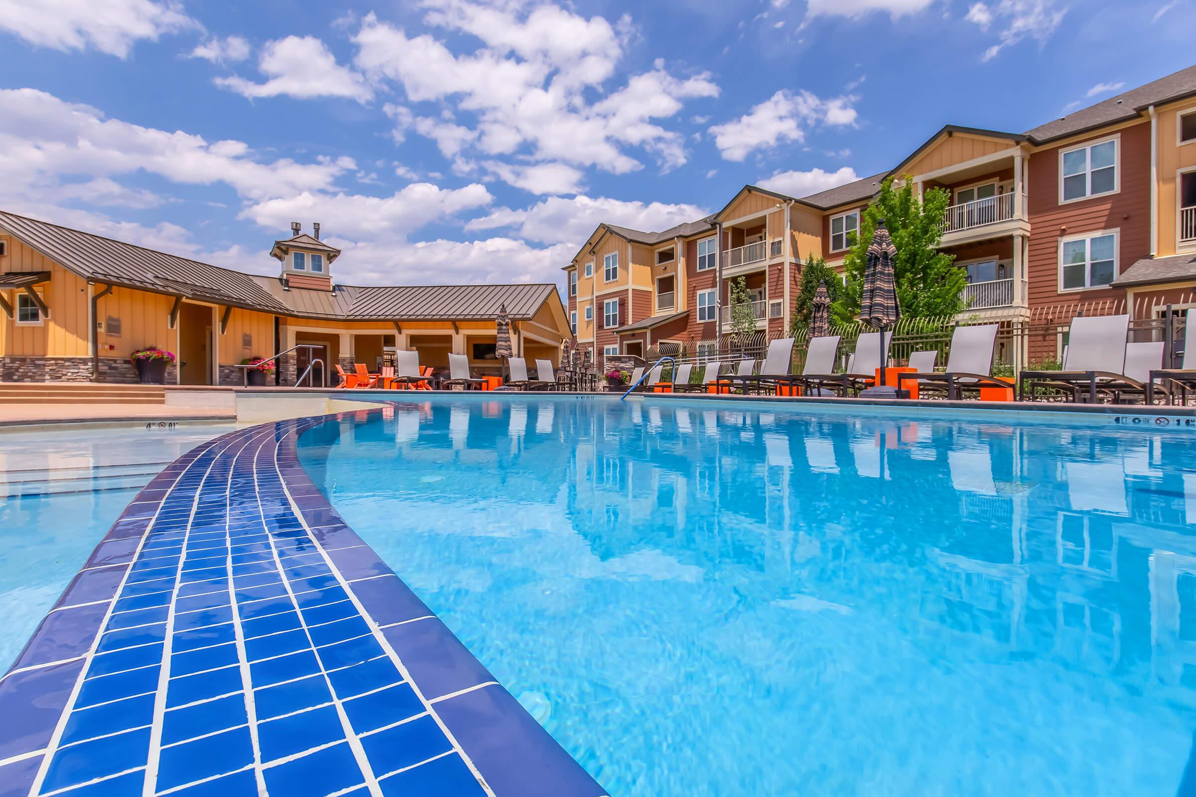 A clear blue swimming pool with a tiled edge, surrounded by lounge chairs and vibrant orange seating. The pool reflects a bright sky filled with fluffy clouds, while a modern building with multiple stories is visible in the background. Green trees add a touch of nature to the setting.