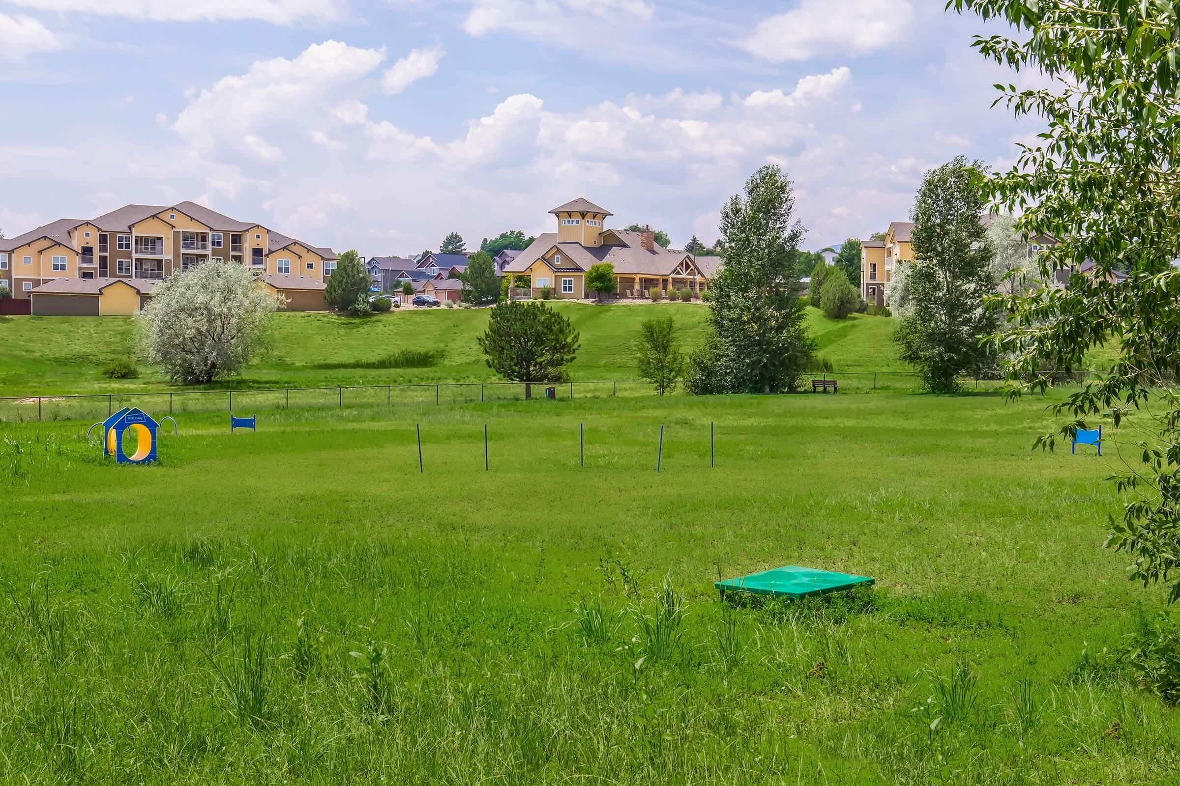 A green grassy field with two colorful play structures on the left, surrounded by trees. In the background, there are several residential buildings and a tall structure with a clock tower under a partly cloudy blue sky.