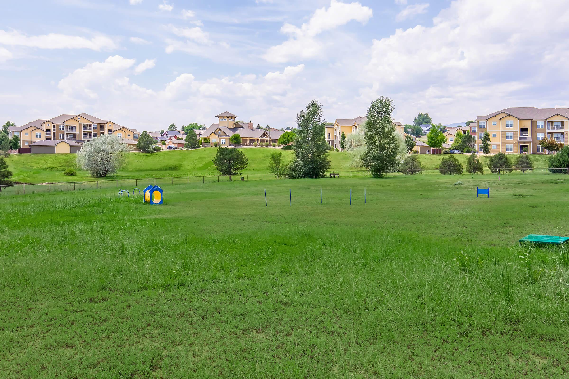 A lush green field stretches across the foreground, dotted with a colorful play structure. In the background, several multi-story residential buildings are situated on a gentle hill, framed by trees under a partly cloudy sky. The scene conveys a peaceful suburban landscape.