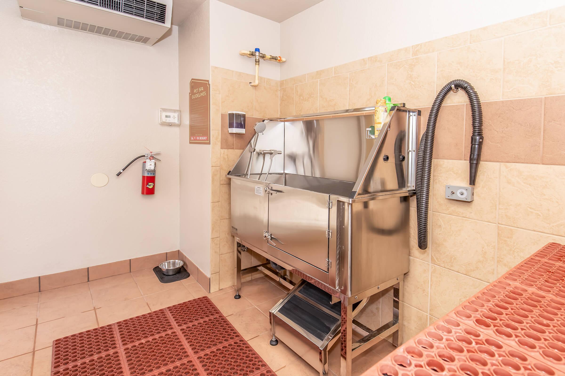 A stainless steel pet washing station in a tiled room. The station features a large tub, a shower hose, and a hose attachment. There's a fire extinguisher on the wall and a small pet bowl on the floor. The area has a mat and soft lighting, creating a welcoming space for pet bathing.