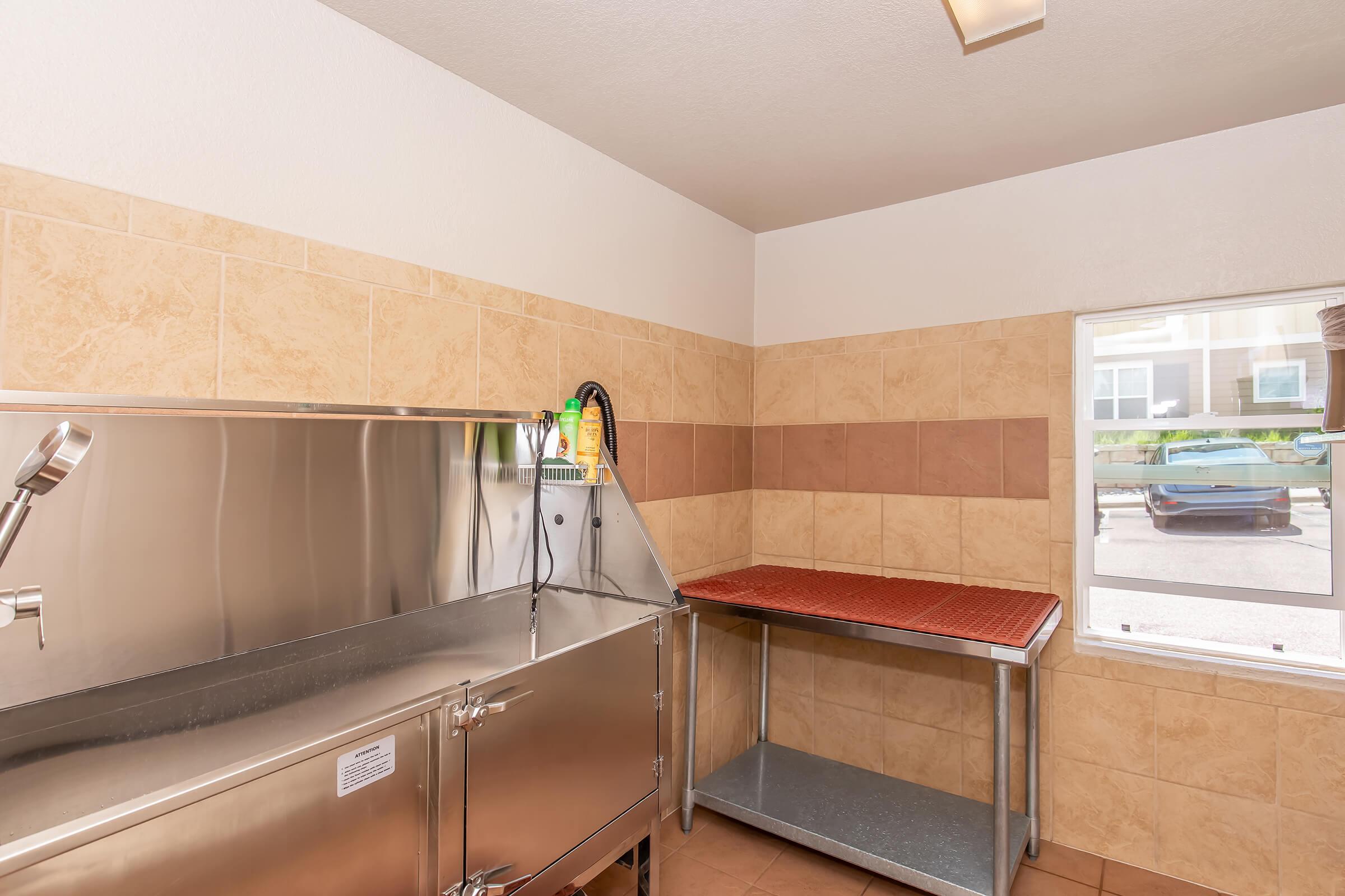 A clean, modern kitchen space featuring a stainless steel sink and countertop. The walls are tiled in a light beige and brown pattern. A window allows natural light to enter, revealing a glimpse of a car parked outside. There is also a metal table on the right side of the room.