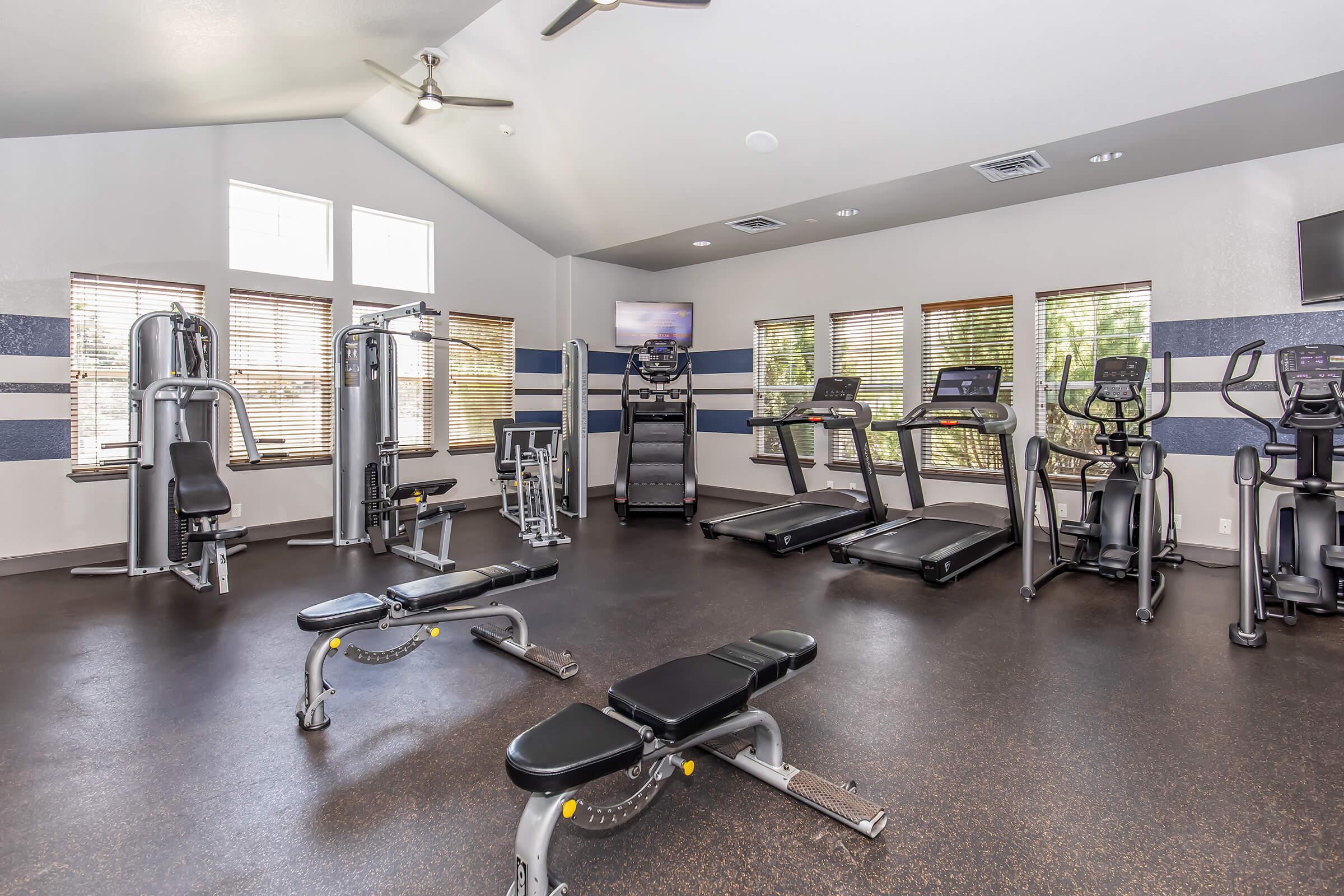 A modern gym interior featuring various fitness equipment, including treadmills, exercise bikes, weight benches, and multi-gym machines. The room has several windows allowing natural light, a ceiling fan for ventilation, and televisions mounted on the walls for entertainment during workouts.