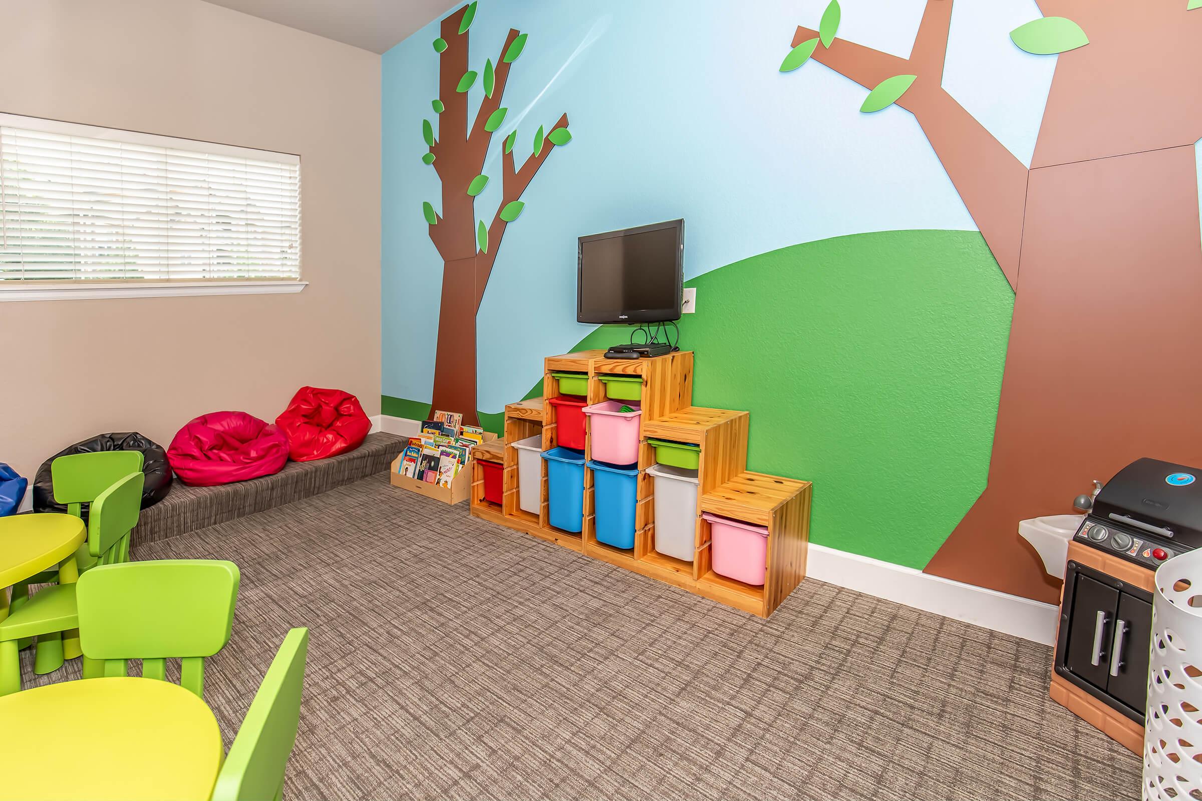 A brightly colored children's playroom featuring green and blue walls with tree decals. There are green tables and chairs, colorful storage bins on a wooden shelf, a TV mounted on the wall, bean bag chairs, and a small play kitchen. The room is designed for play and creativity.