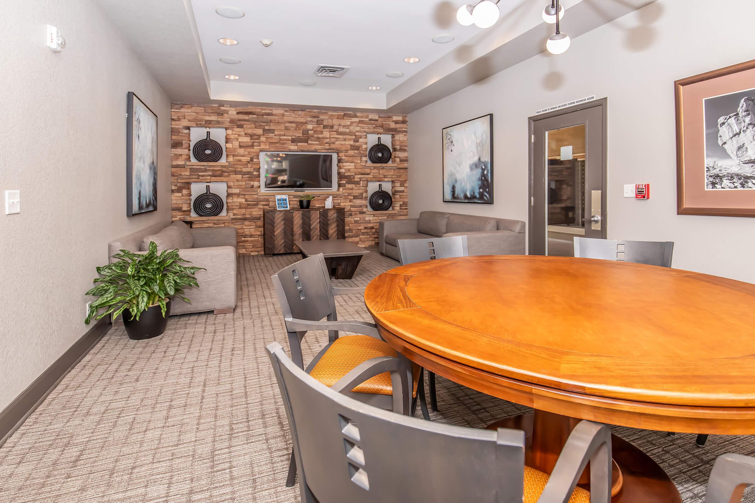 A modern lounge area featuring a round wooden table surrounded by black chairs, with comfortable gray sofas in the background. A wood-paneled wall holds a flat-screen TV and speakers. Ambient lighting from ceiling fixtures creates a cozy atmosphere. A potted plant adds a touch of greenery to the space.
