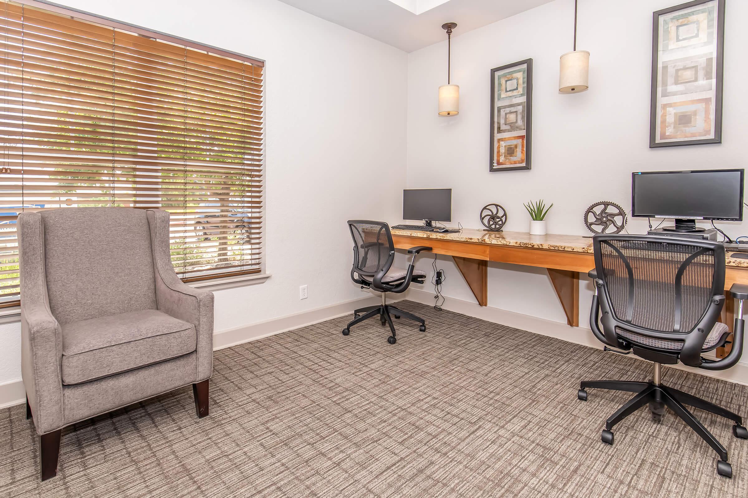 A modern workspace featuring two computer desks with monitors, a comfortable armchair, and decorative wall art. Bright window with blinds allows natural light, complementing the neutral color palette and contemporary design of the room.