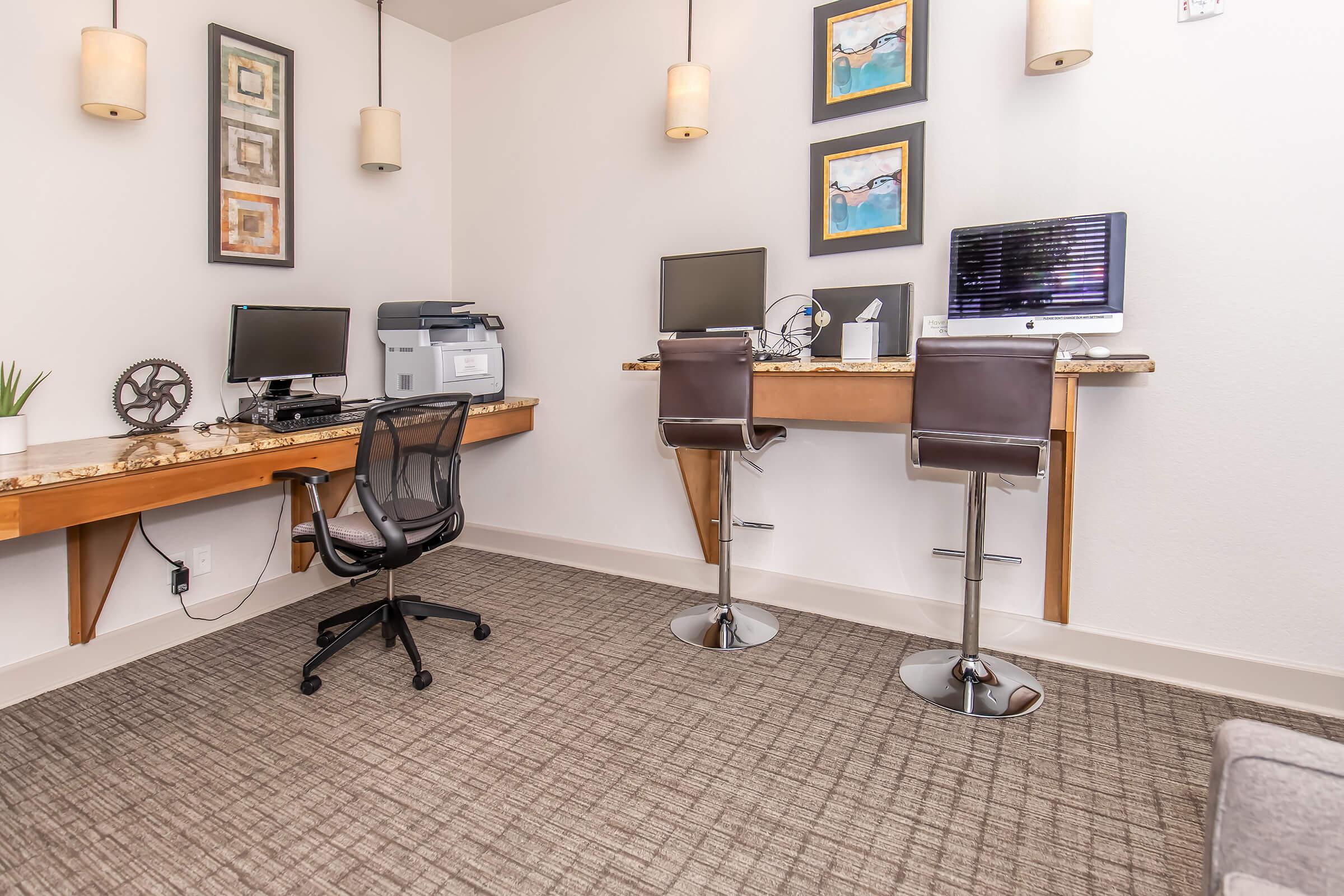A modern workspace featuring two desks with computers, a printer, and stylish bar stools. The walls are adorned with framed wall art, and the floor is covered with a neutral-toned carpet, creating a professional and inviting atmosphere.