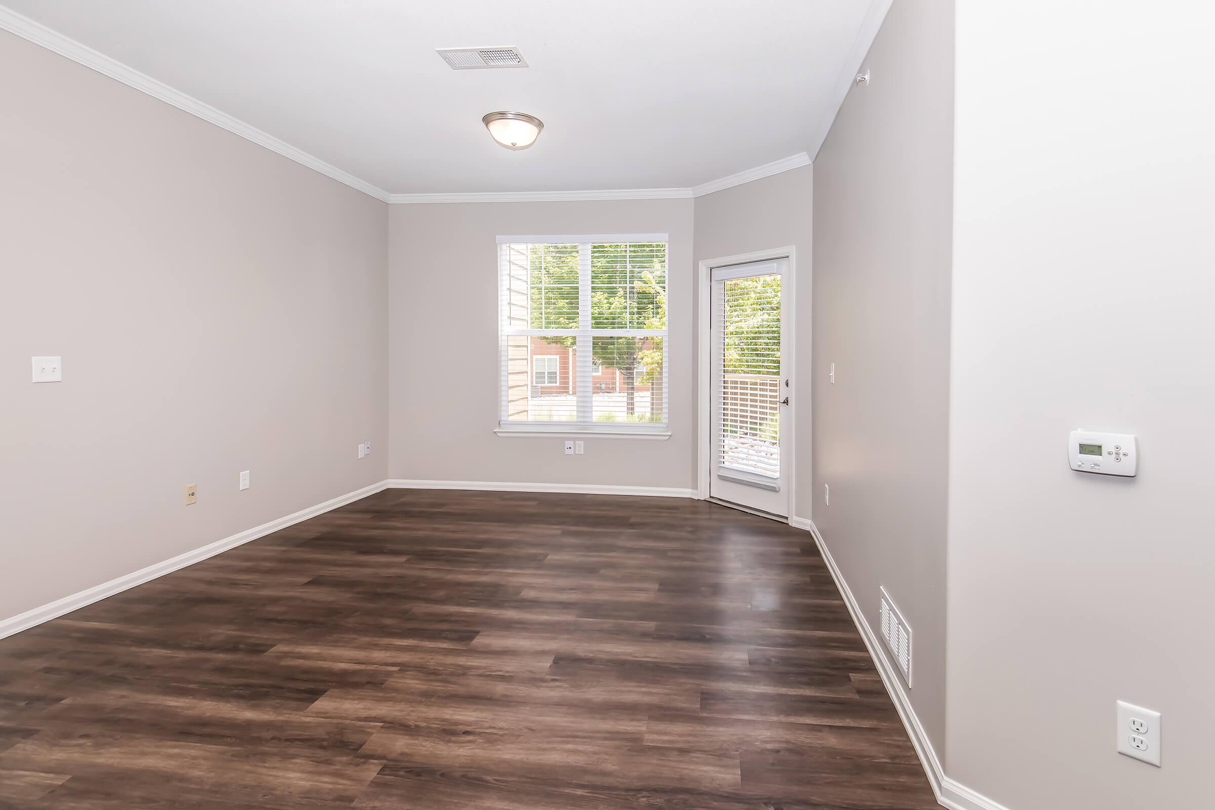 Empty interior of a room with light gray walls and dark wooden flooring. A single window with white blinds allows natural light in, and there is a door leading outside. The space includes a ceiling light fixture and a thermostat on the wall.