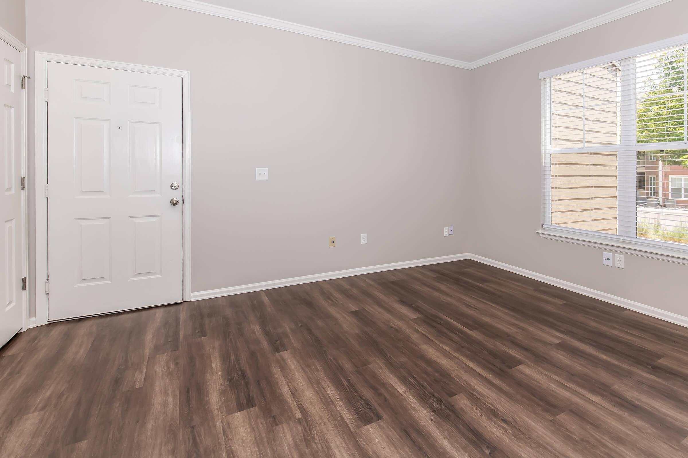 An empty room with light gray walls and dark wood-like flooring. There is a white door on the left side and a large window on the right, allowing natural light to enter. The room has no furniture, creating a spacious and open appearance.