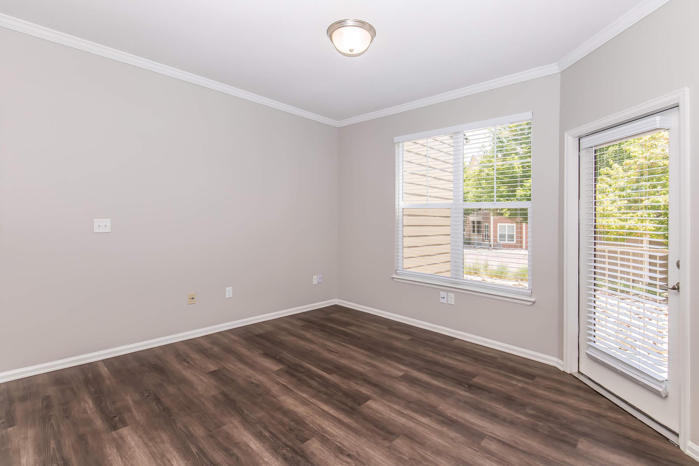 Empty interior of a room with light gray walls and a window featuring white blinds. The floor is dark wood laminate, and there's a door leading outside. A ceiling light fixture is present, adding brightness to the space. The overall atmosphere is clean and minimalist, ideal for various uses.