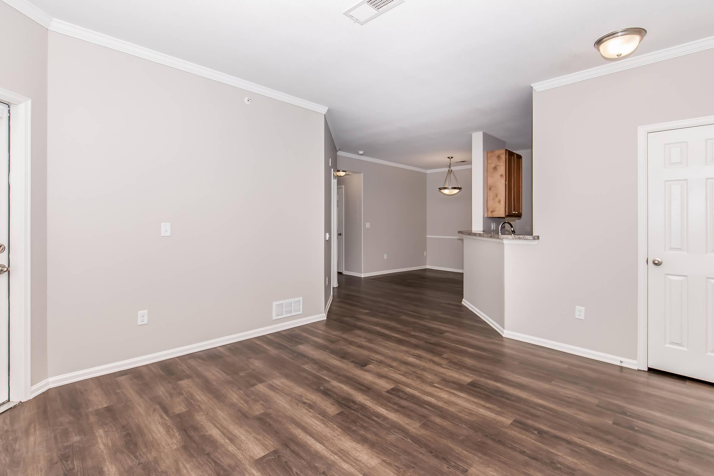 A spacious, neutral-colored living area with laminate flooring and a smooth ceiling. On the left, a doorway leads to the entrance, while a partial wall opens to a kitchen area in the background. The kitchen features wood cabinetry visible from the living space. Natural light fills the room.