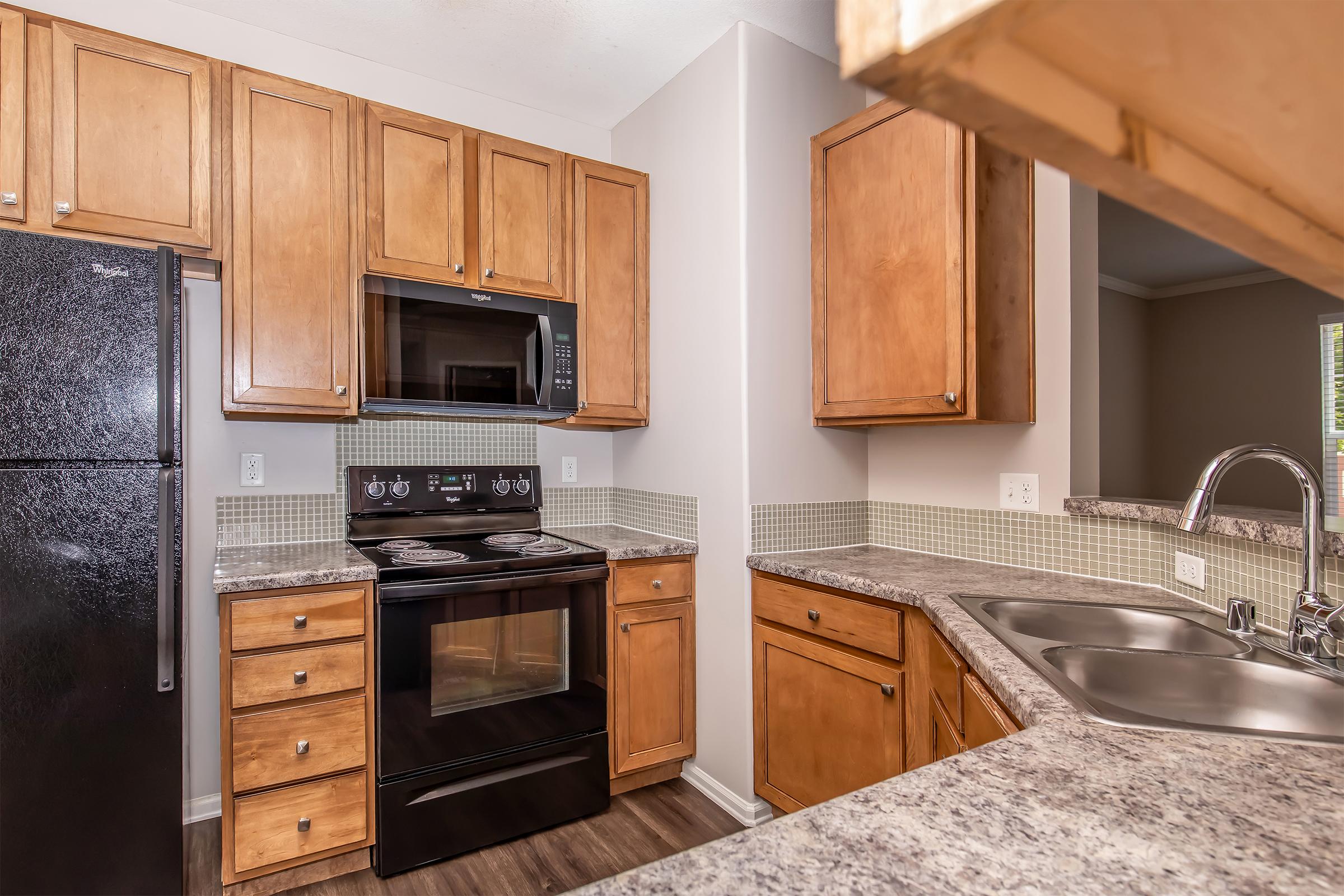 A modern kitchen featuring wooden cabinets, a black refrigerator, and an oven with a stovetop. The countertops are made of gray granite, and there's a double sink with a pull-down faucet. The walls are painted a neutral color, and a mosaic tile backsplash adds a decorative touch.