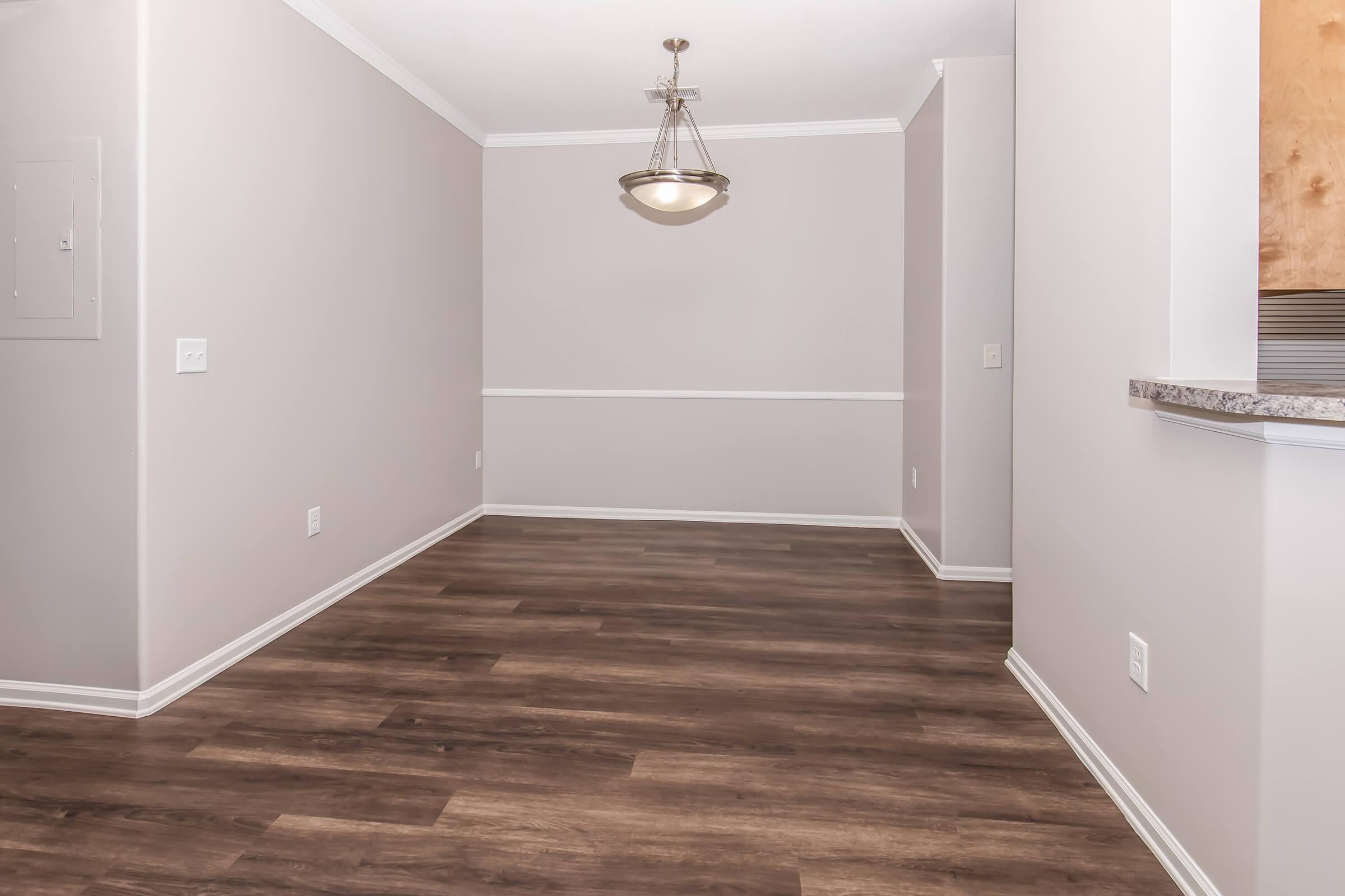 A spacious and well-lit room featuring light beige walls, elegant crown molding, and dark wooden flooring. A modern chandelier hangs from the ceiling, illuminating the area. The room is empty, highlighting its potential for various uses, with a partial view of a kitchen on the right side.