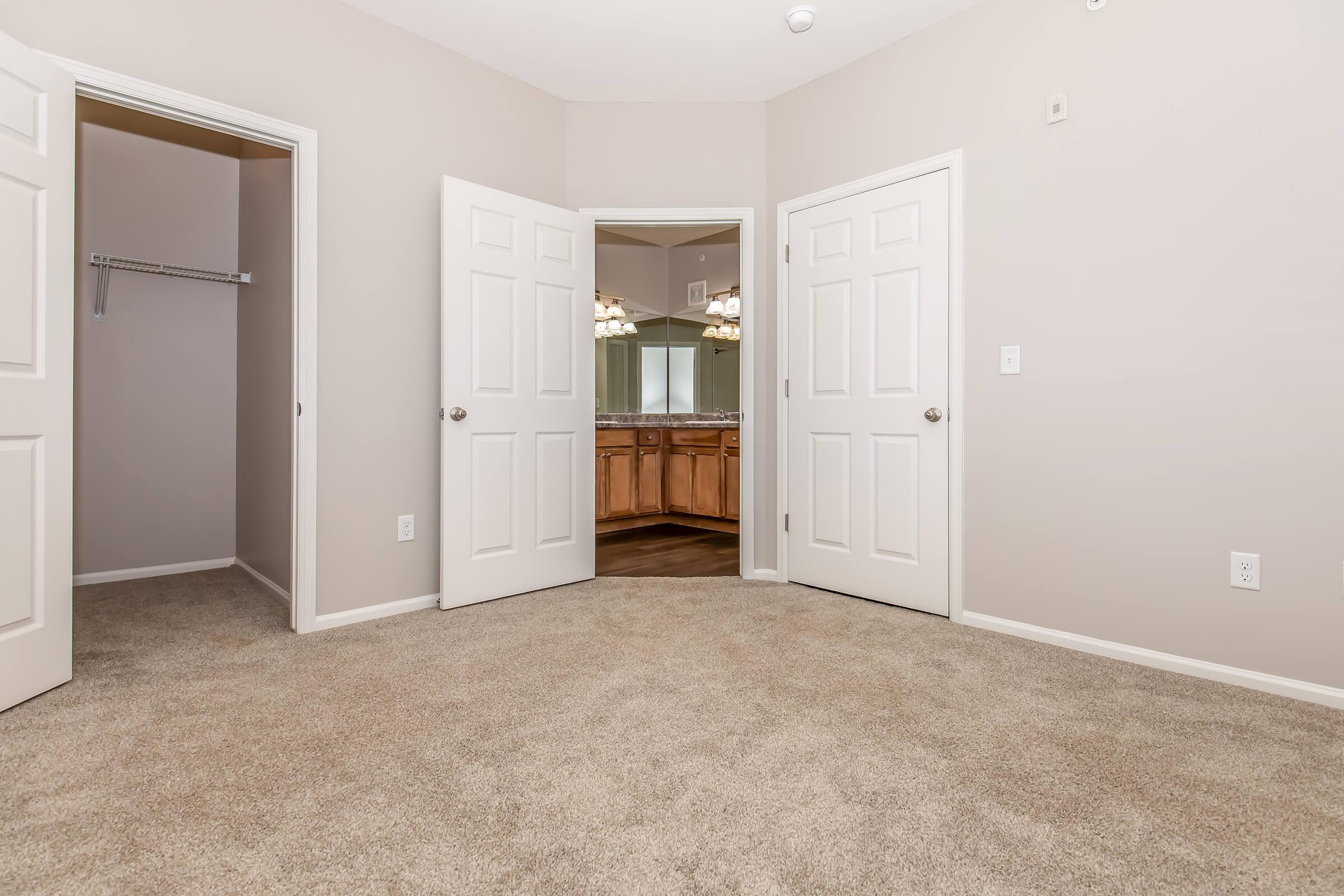 A beige room featuring plush carpet, two open doors leading to a closet and a bathroom area. The bathroom can be seen in the background with wooden cabinetry and lighting fixtures. Soft natural light fills the space, creating a cozy atmosphere.