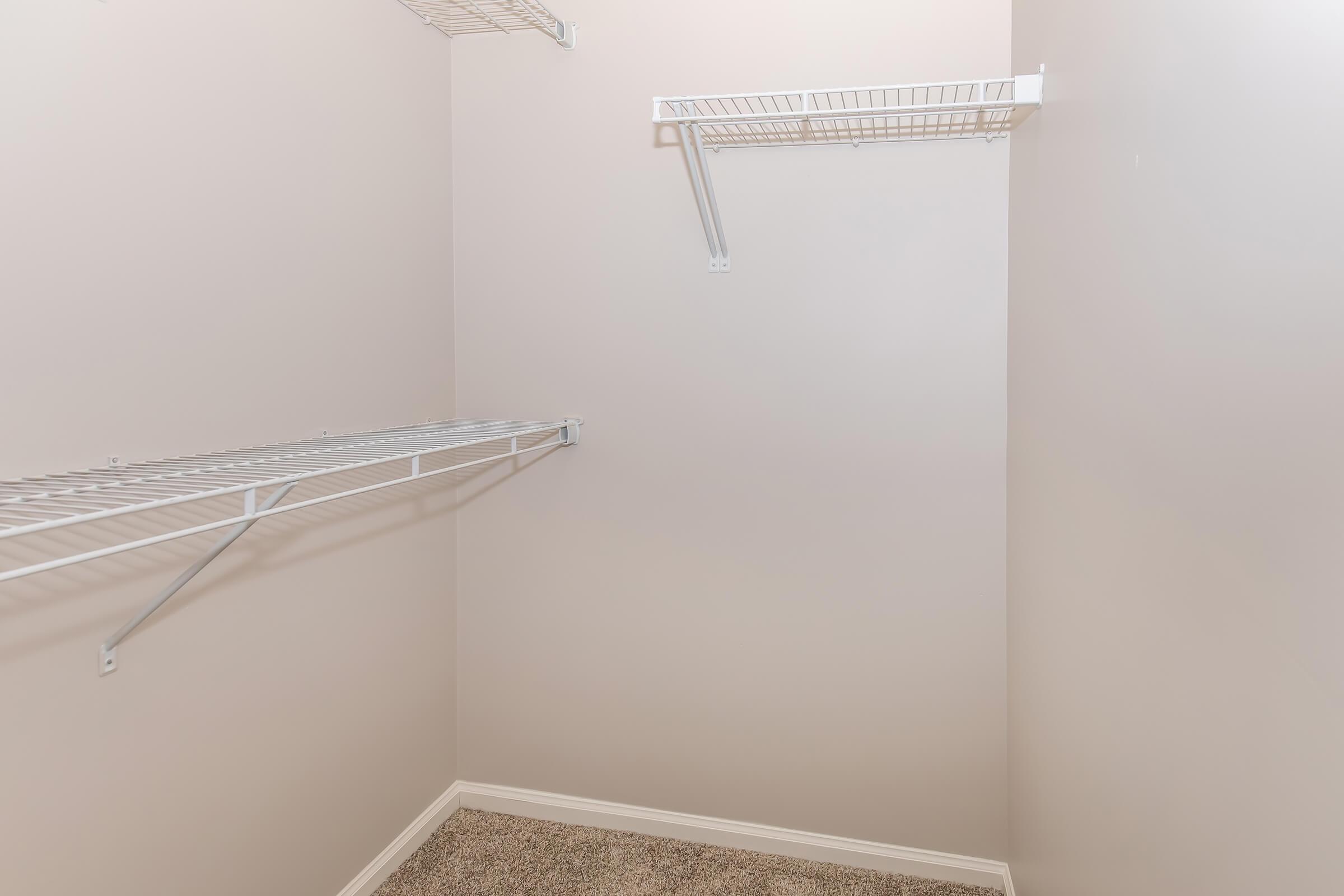 Empty closet with light-colored walls. It features two metal wire shelving units mounted on the walls, showing a clean and organized space. The floor is covered in a light carpet, enhancing the bright and airy feel of the area.