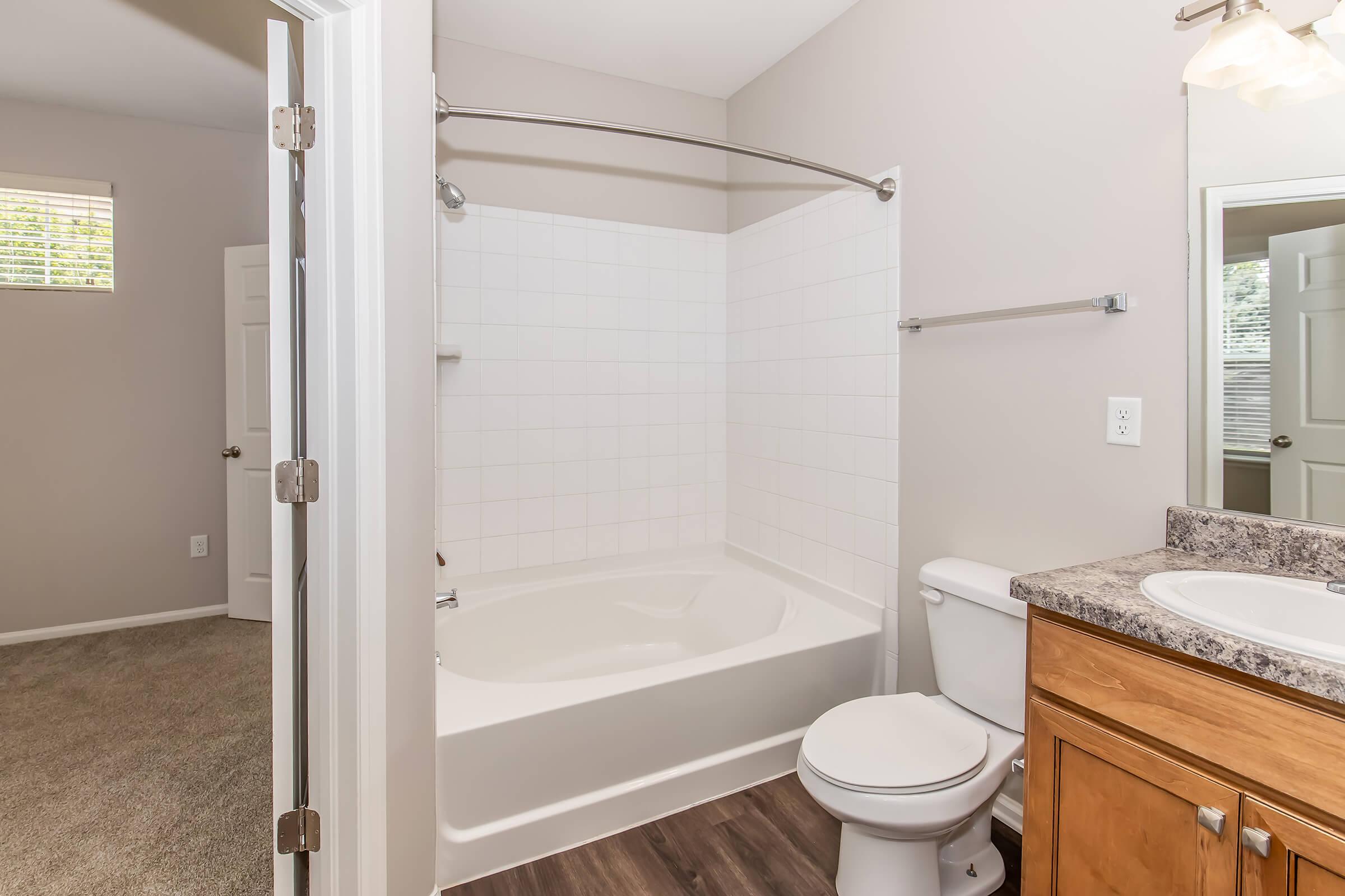 A clean bathroom featuring a bathtub with a shower, a toilet, and a vanity with a sink. The walls are painted in a light color, and there's a window allowing natural light. The floor has a wood-like appearance, and the bathroom is well-organized and tidy, showcasing a modern aesthetic.