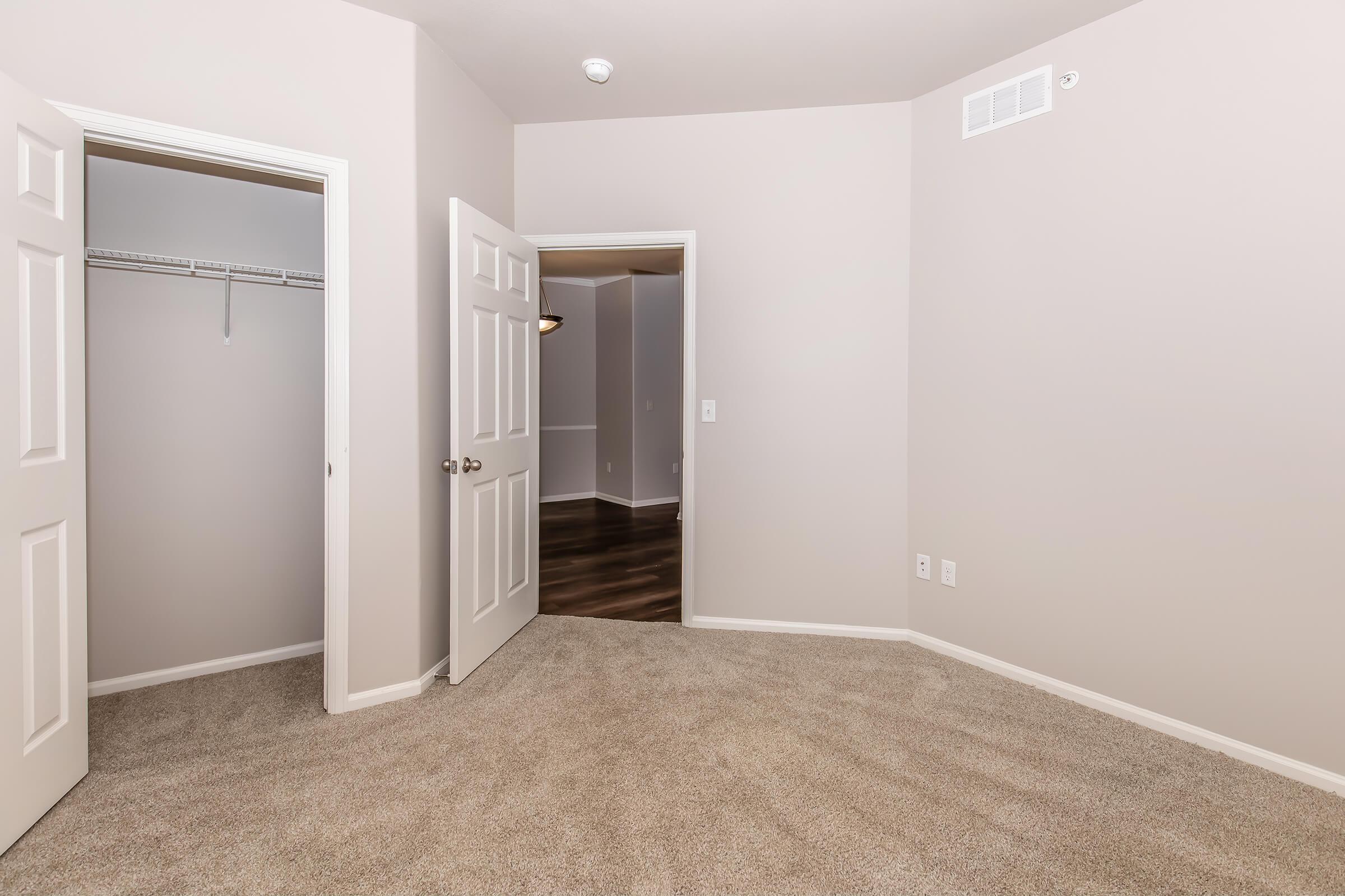 Empty room featuring light-colored walls and carpet. There are two doors: one leading to a closet and another potentially leading to another area. The floor is wood-like, and the space is well-lit, giving it an open and airy feel.