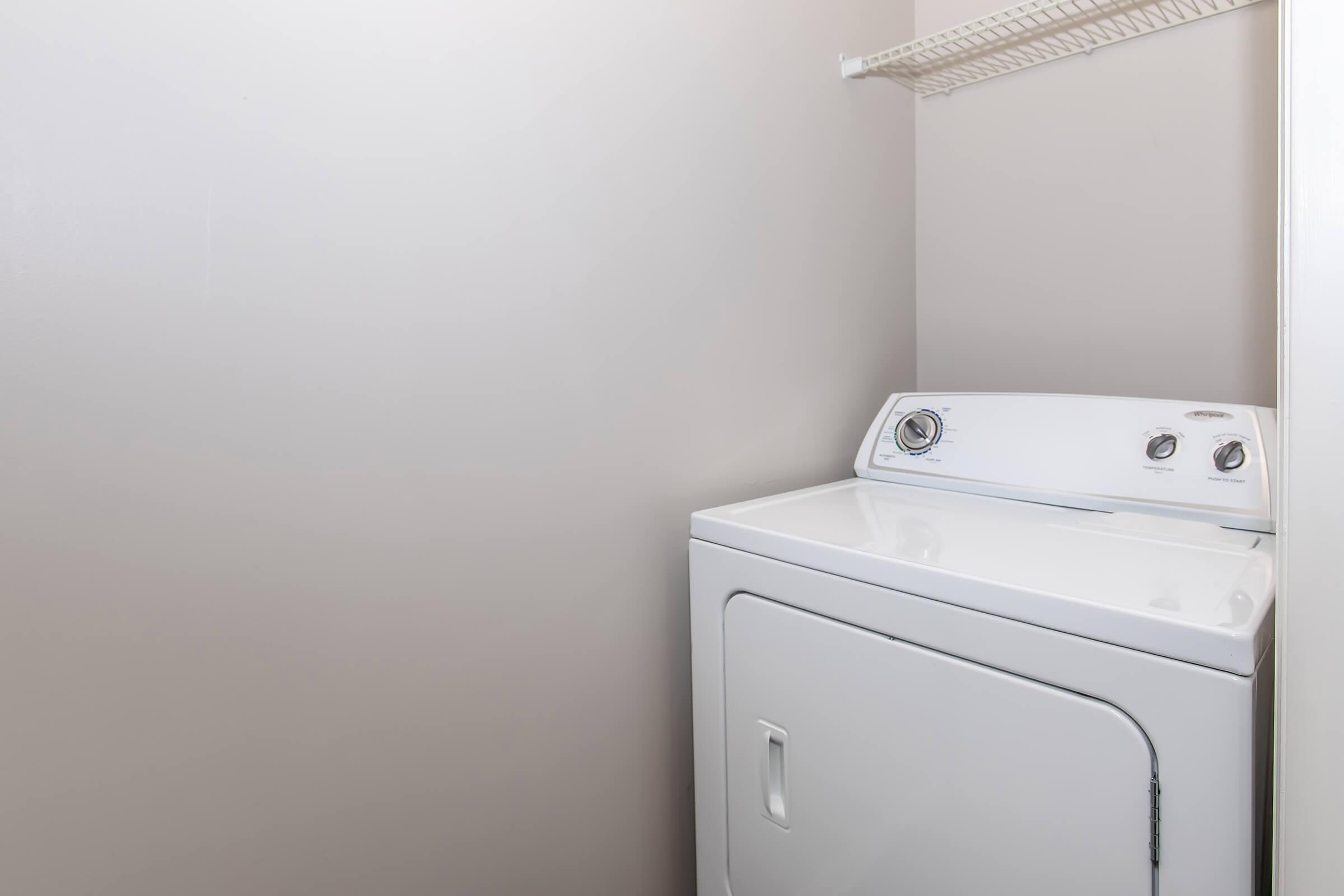 A clean, white washing machine situated in a corner of a laundry room. The wall is painted light gray, and there is a wire shelf above the machine, providing storage space. The overall setting is tidy and minimalistic.