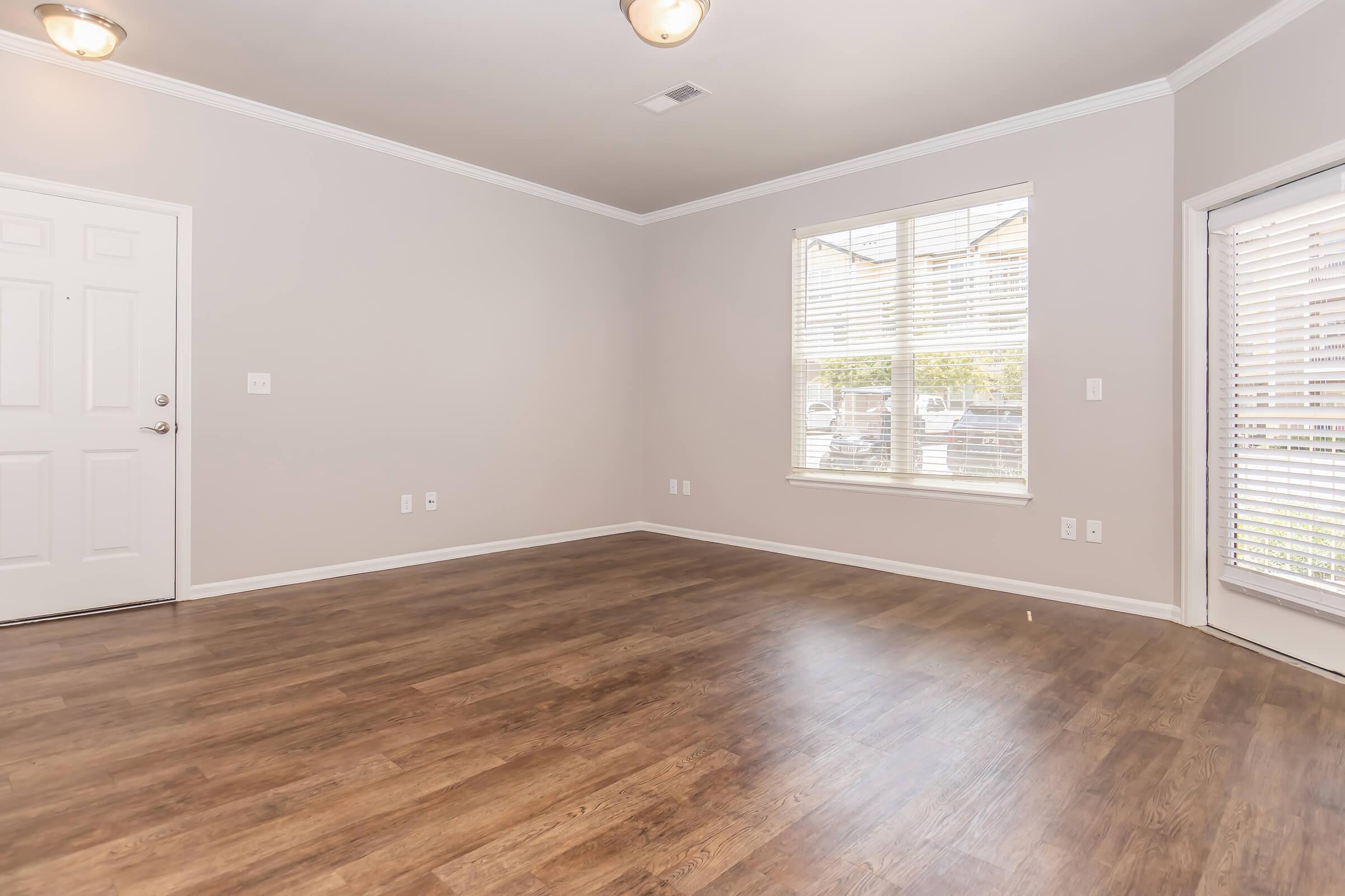Empty room with light-colored walls and a brown wooden floor. One wall features a door and a window with blinds, allowing natural light to enter. Another wall has a large window with horizontal blinds. The space appears clean and well-lit, ideal for various interior designs.