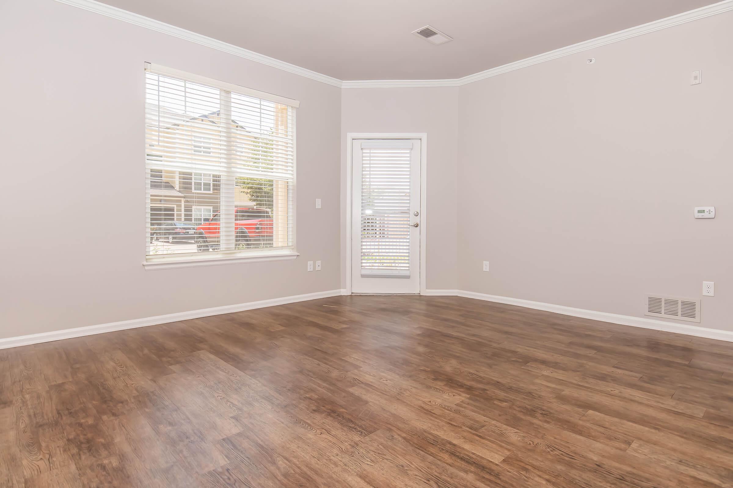 A spacious, empty room with light-colored walls and a wooden floor. There are large windows allowing natural light, and a door leading outside. The room has baseboards and a ceiling crown molding, creating a bright and inviting atmosphere.