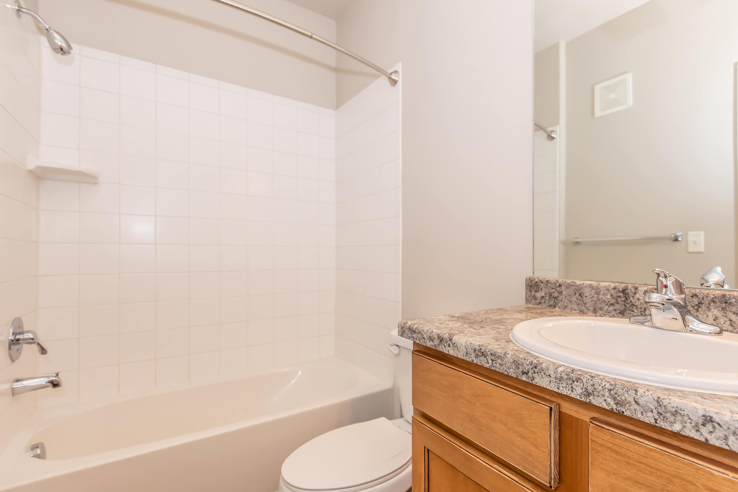 A clean bathroom featuring a bath/shower combo, a granite countertop with a sink, and wooden cabinetry. The walls are light-colored, and there is a towel bar visible in the background. The space is well-lit and tidy, suggesting a modern and functional design.