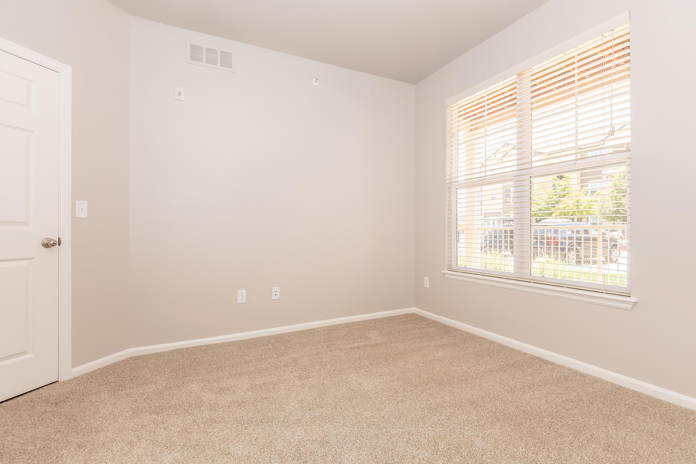 Empty room with light gray walls and beige carpet. A large window with white blinds lets in natural light, overlooking an outdoor area. A single door is visible on the left side of the image. The room appears clean and well-maintained, ideal for use as a bedroom or office space.