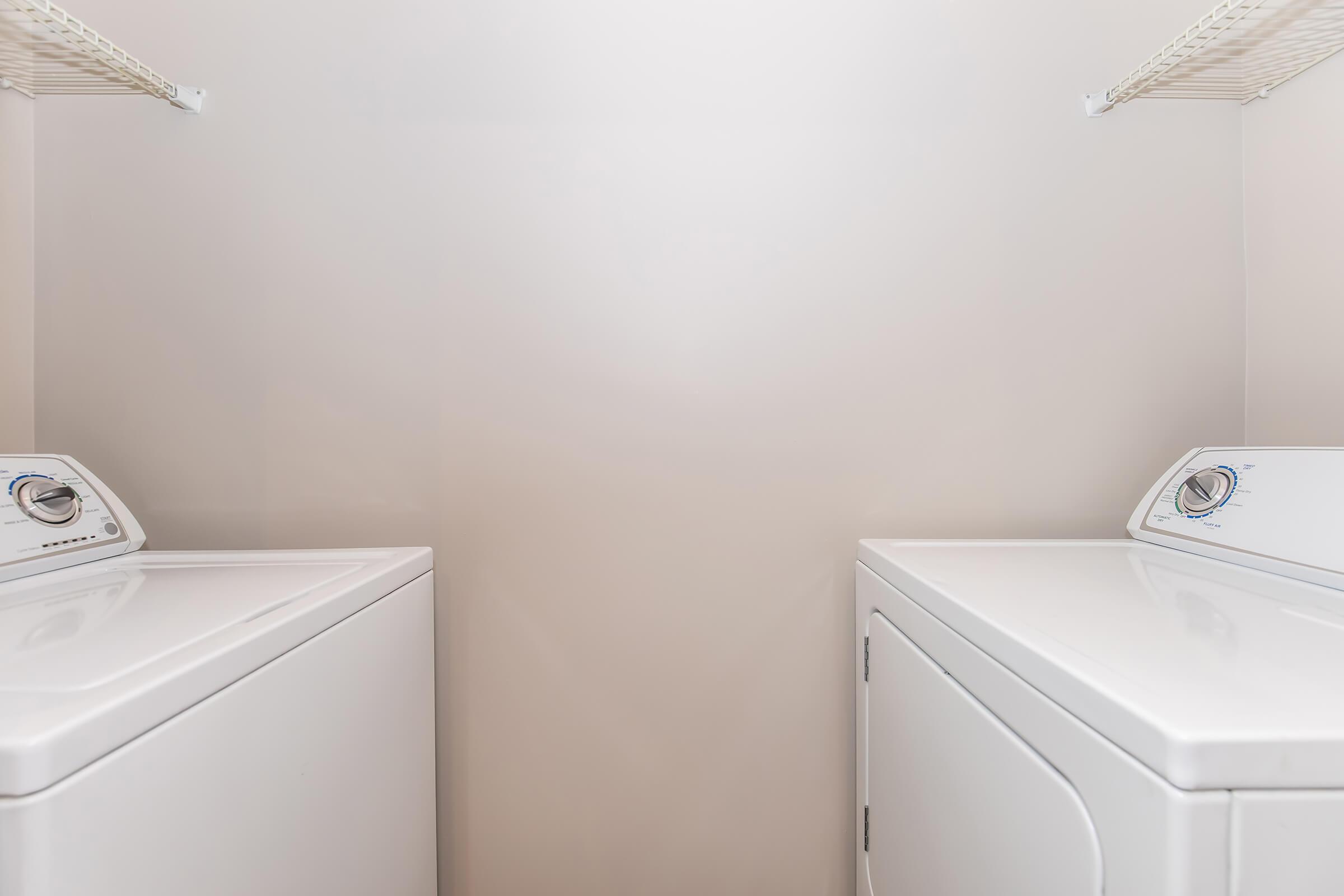 Two white laundry appliances, a washer and a dryer, are positioned side by side against a light-colored wall. The shelves above are empty, and the space appears clean and organized, suitable for laundry tasks.